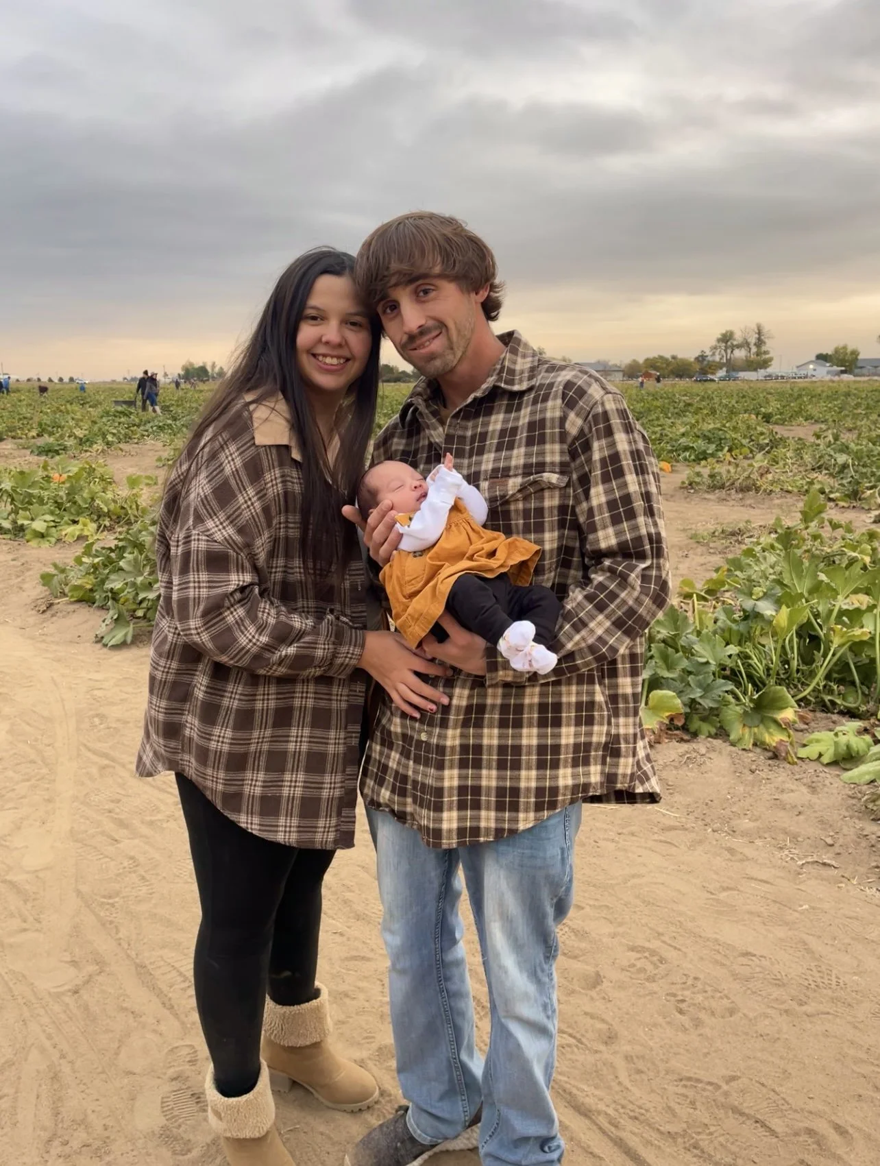 A mother and father stand together in a pumpkin patch, smiling and holding their newborn baby dressed in fall colors.