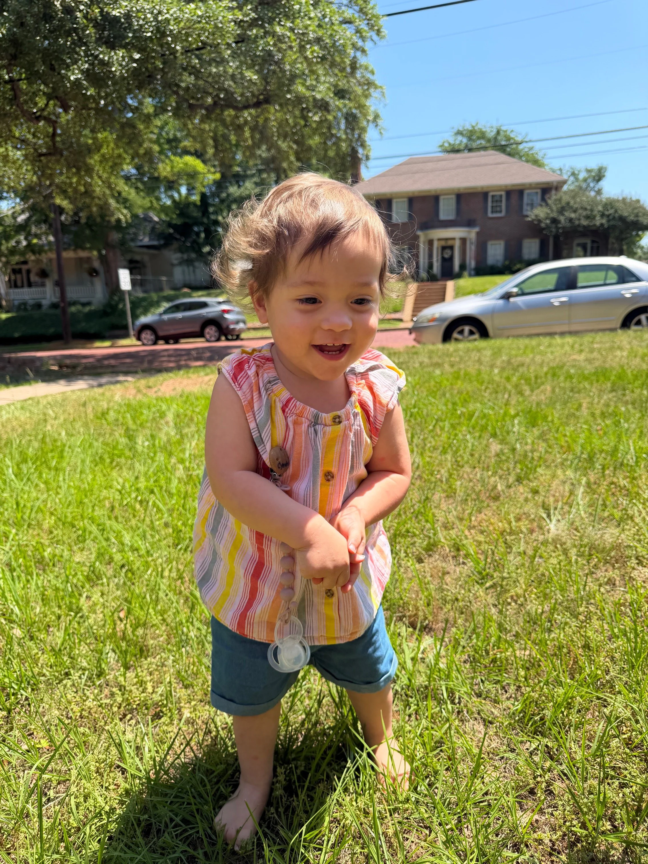 Smiling toddler in colorful shirt stands barefoot on grass in a sunny yard with houses and cars in the background.