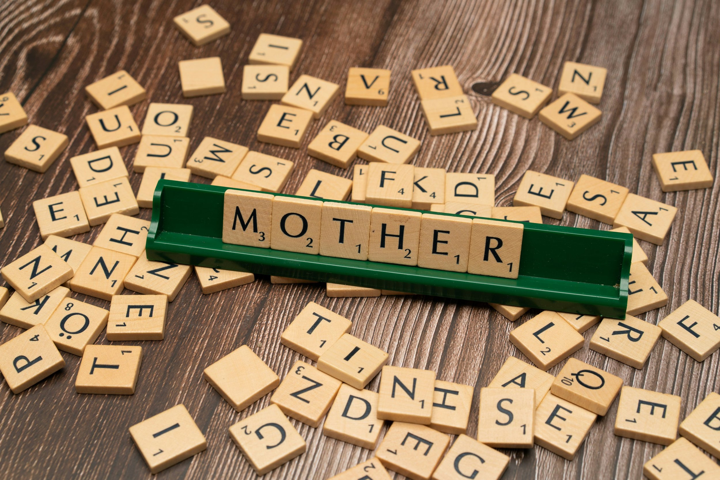Photo of light wooden letter tiles spelling "MOTHER" on a dark wooden background scattered with other letter tiles.