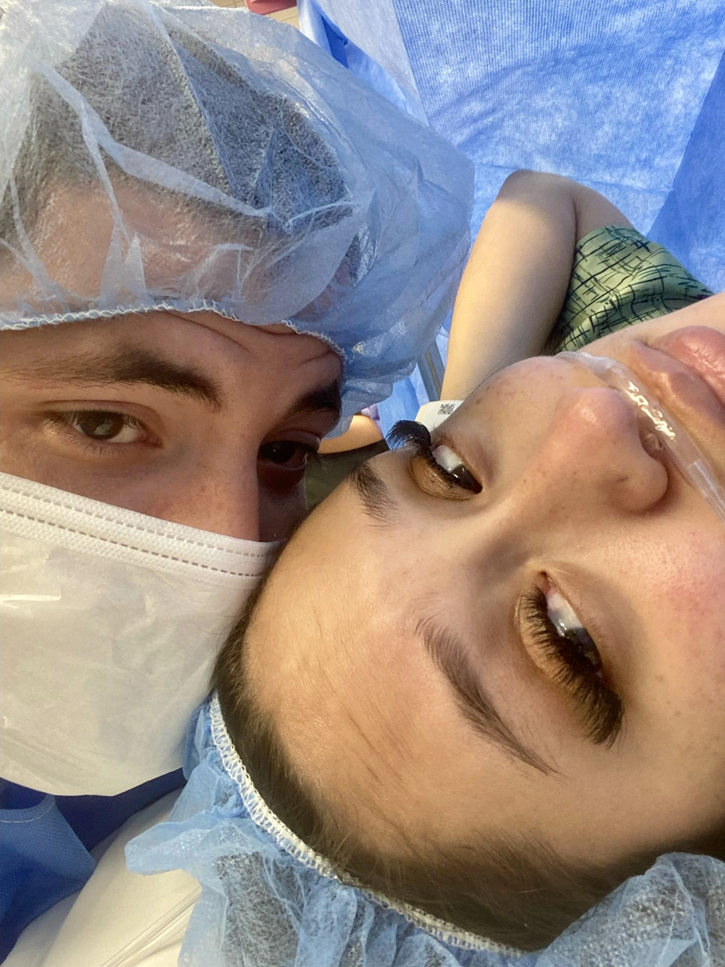 Two people in medical caps and gowns lie close together, one wearing an oxygen tube, both looking at the camera.