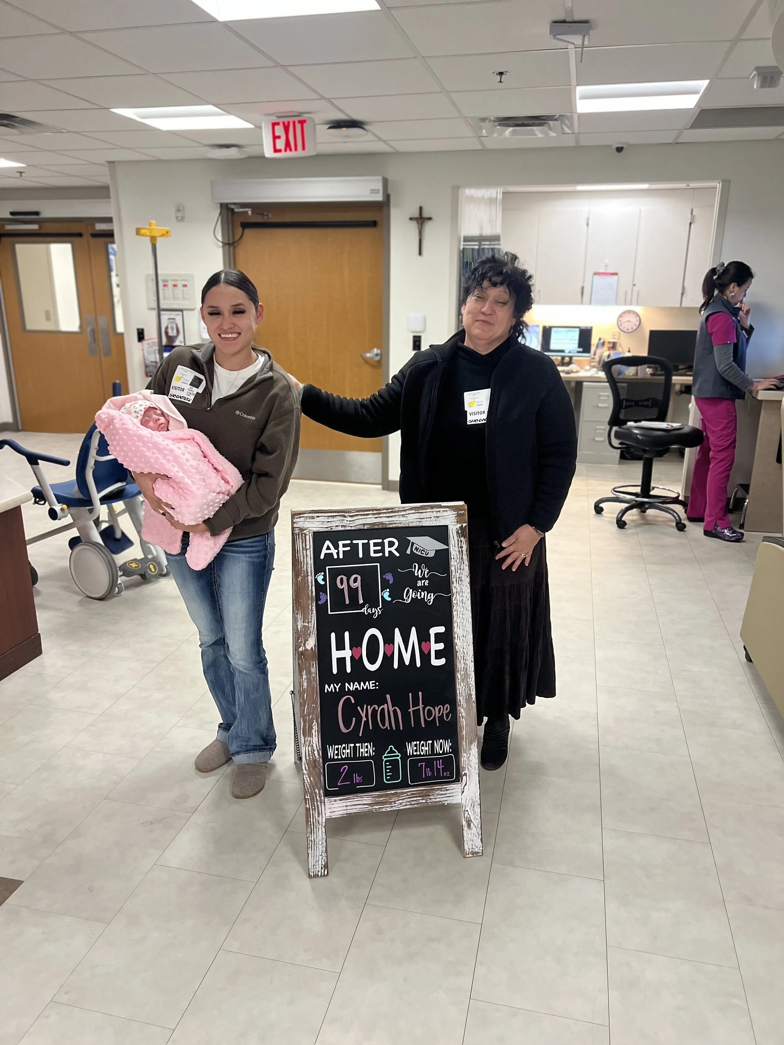 Two women stand in a hospital next to a chalkboard sign celebrating a newborn named Cyrah Hope going home.