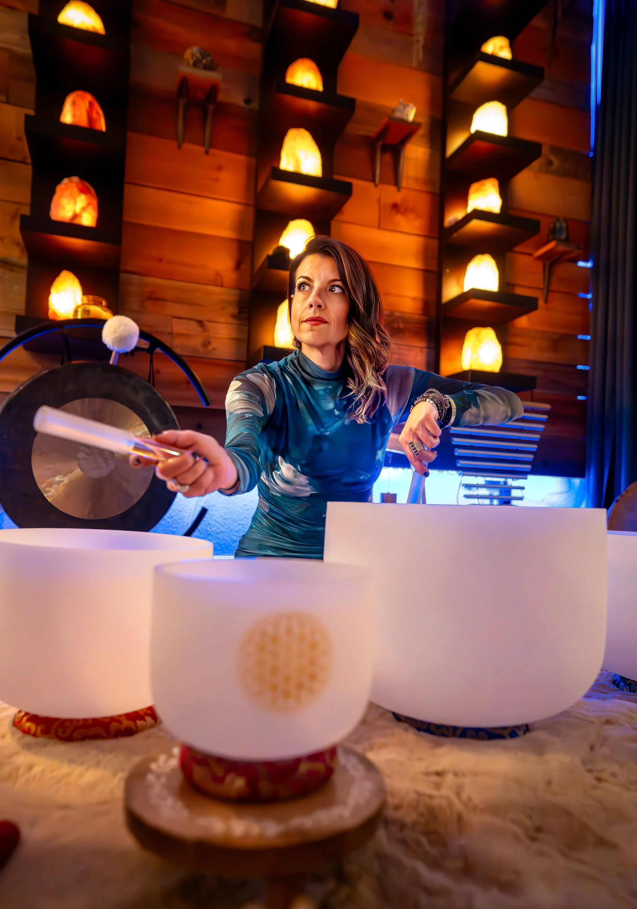 Woman sitting at a table with two white bowls in the foreground, decorated with red and gold patterns, in a warmly lit restaurant with salt lamps on wooden wall shelves in the background.