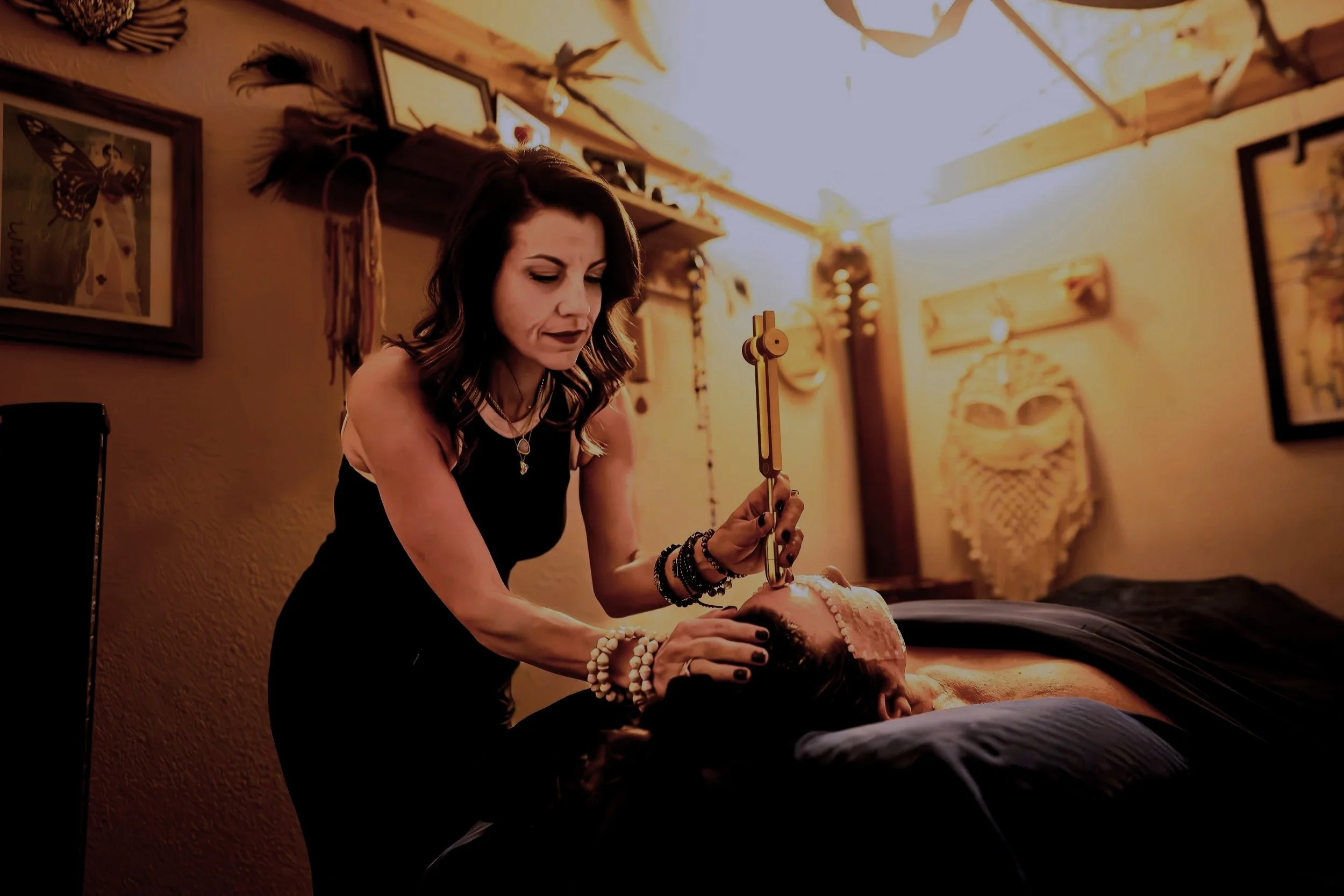 A woman performing a healing or massage on a person lying on a table in a warmly lit room decorated with artwork and plants.