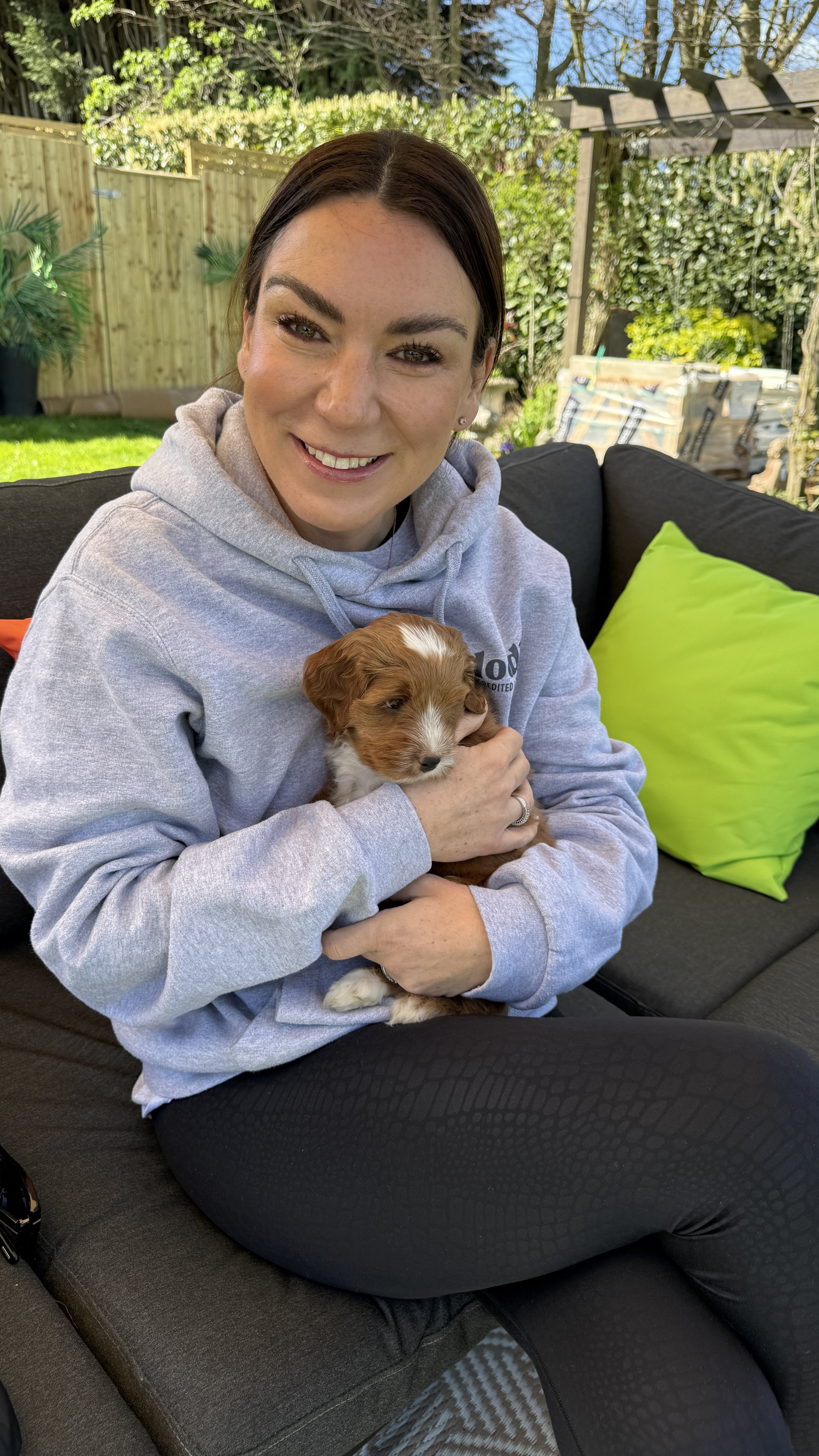 A woman sitting on a black outdoor sofa holding a small brown and white puppy, with a green cushion placed beside her. She is smiling and wearing a light grey hoodie and black leggings. The background shows a garden with trees, a wooden fence, and some garden supplies.
