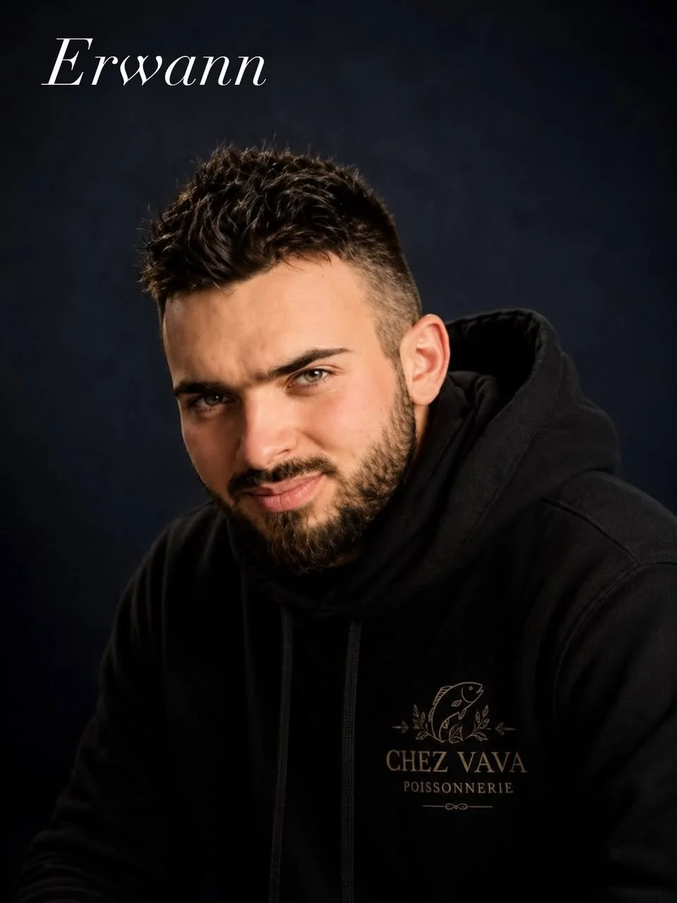 Portrait d'un jeune homme aux cheveux foncés, barbe épaisse, portant un sweat noir avec un logo de poisson et le texte "CHEZ VAVA POISSONNERIE" sur la poitrine, contre un fond sombre.