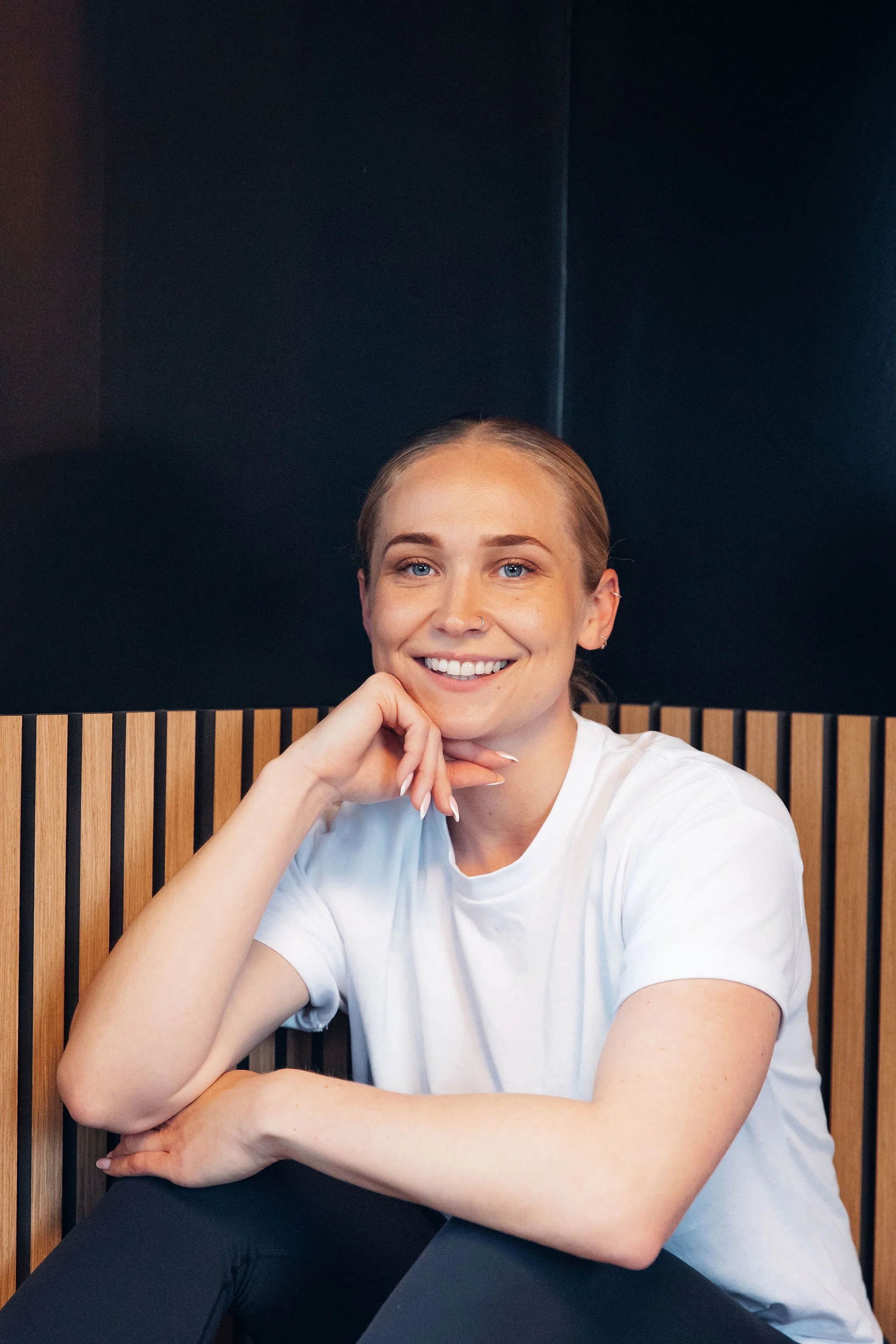 A young woman with blonde hair, blue eyes, and a nose piercing, smiling while sitting with her hand resting on her chin and her arm crossed on her lap, against a black wall and striped wooden panel background.