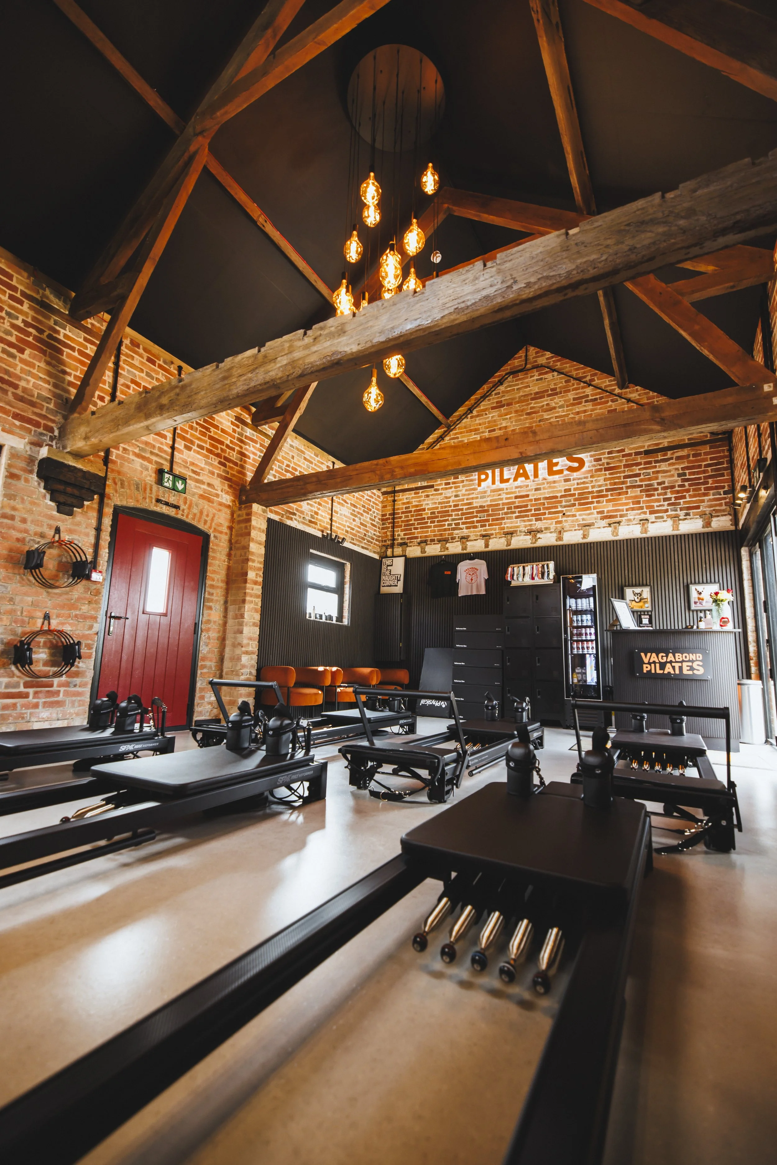 Interior of a Pilates studio with brick walls and exposed wooden beams on the ceiling. Pilates reformer machines are arranged in rows, and a sign that reads 'PILATES' is on the wall. There is a red door and decorative elements on the walls, including shelves with clothing and artwork.