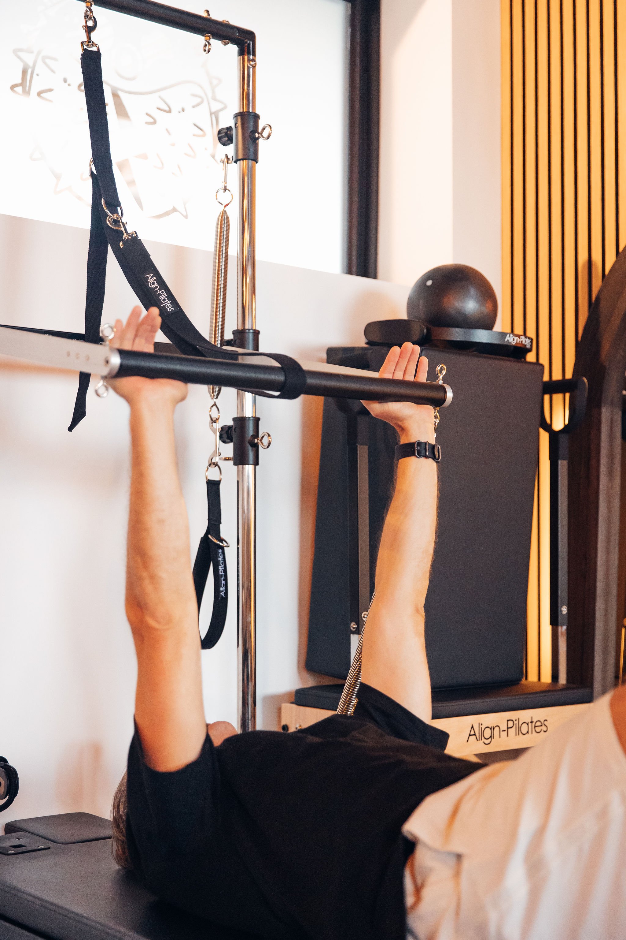 Person lying on Pilates tower and cadillac during a Pilates workout session, with hands gripping the bar above their head, in a studio with wooden wall accents.