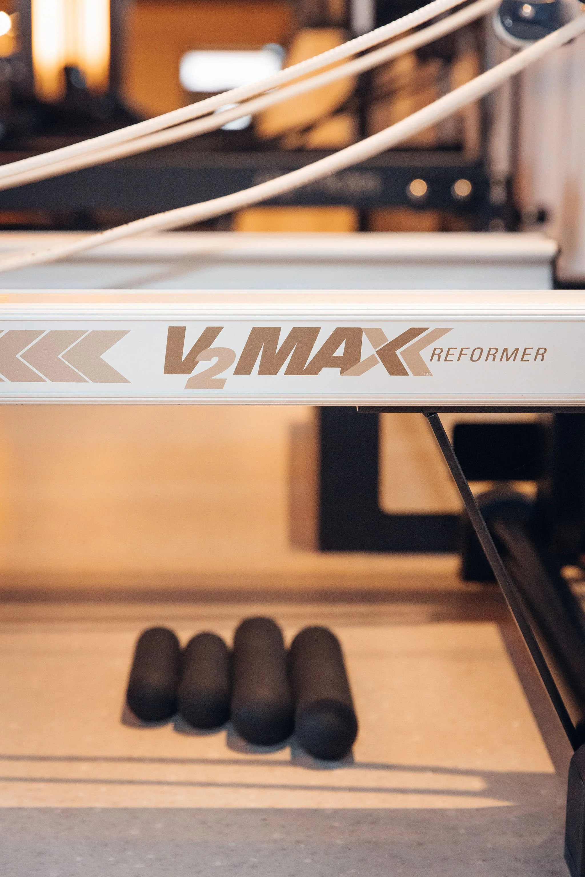 The image shows a ping pong table with a partial logo that reads 'V2MAX REFORMER'. Below the table, there are four black foam rollers aligned on the floor. The background is blurred, indicating an indoor setting.