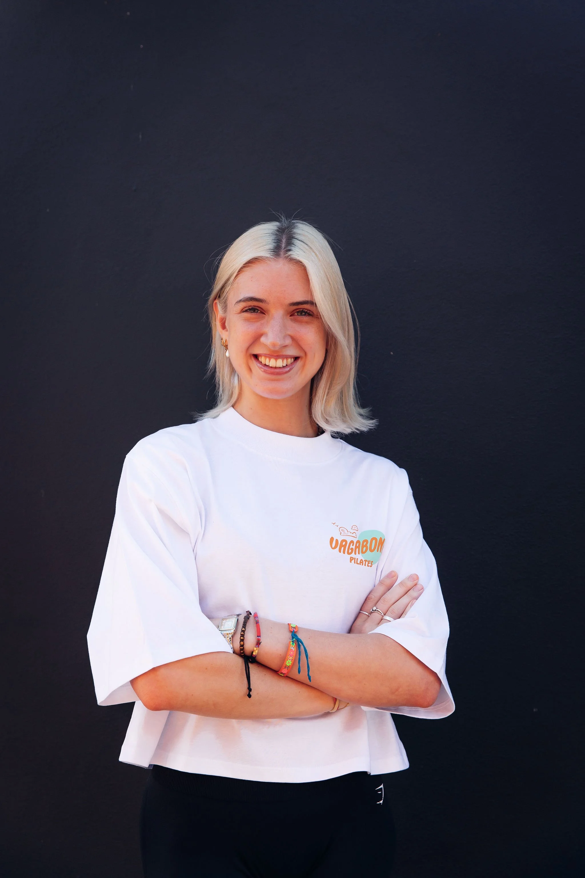 A woman with blonde hair and light skin standing in front of a dark background, smiling with crossed arms, wearing a white t-shirt with an orange and white logo that reads "Uagabon Pilates," and colorful bracelets on her wrist.