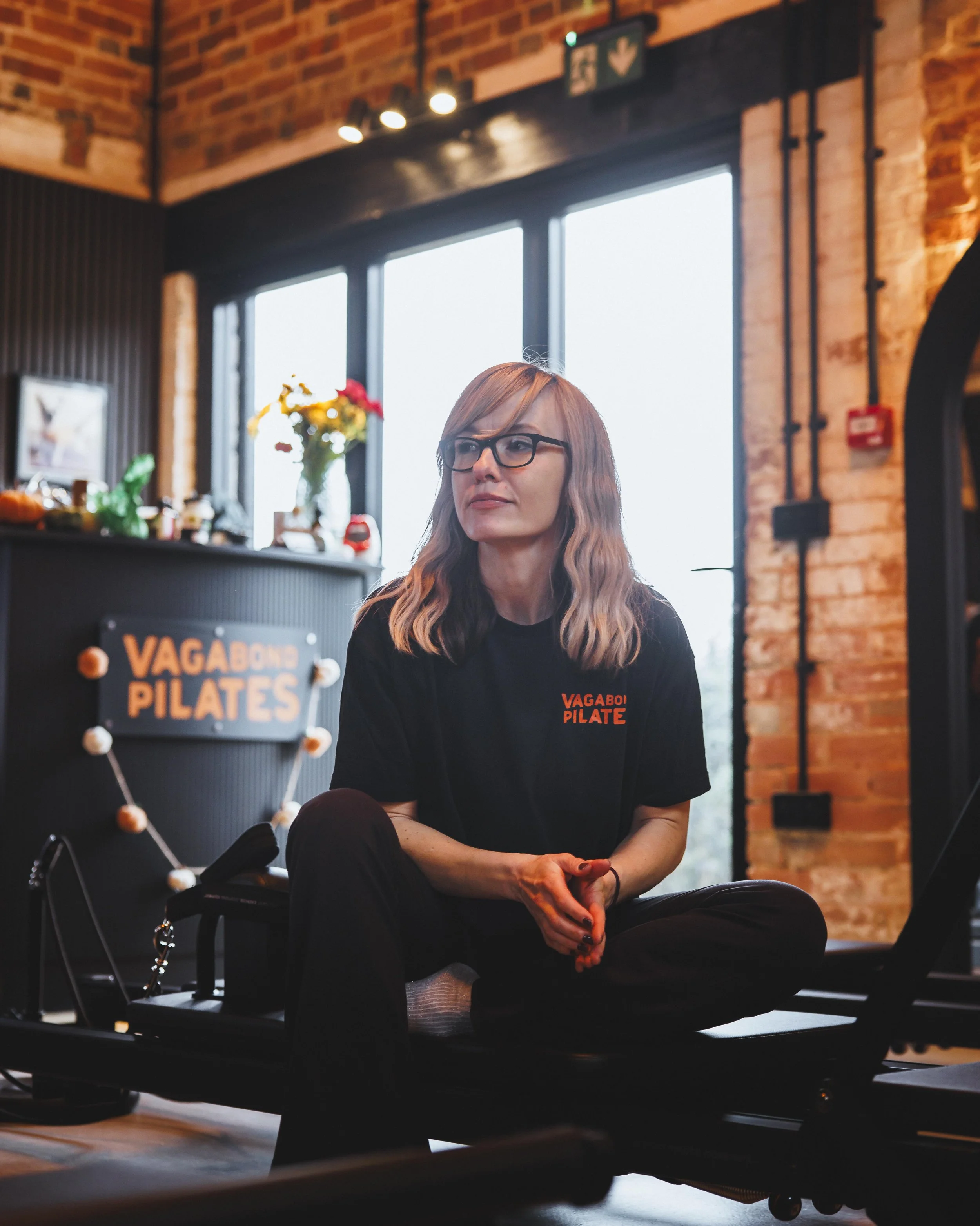 A woman with glasses and wavy blonde hair sitting cross-legged on a Pilates reformer machine in a fitness studio, wearing a black T-shirt that says 'VAGABOND PILATES.' The studio has exposed brick walls, large windows, and a sign in the background that also says 'VAGABOND PILATES.'