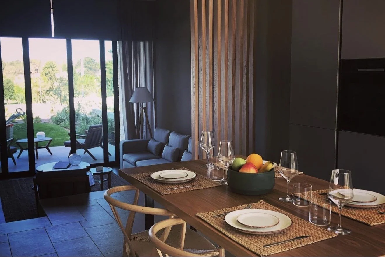 Dining table with plates, glasses, and a bowl of fruit in a modern living space with a view of an outdoor patio and garden.