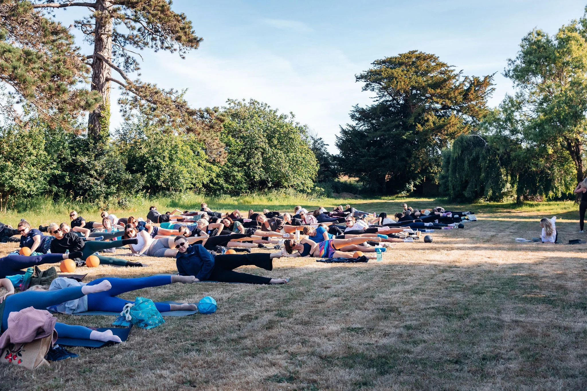 Group of people participating in an outdoor yoga or stretching class on a grassy field surrounded by trees on a clear day.