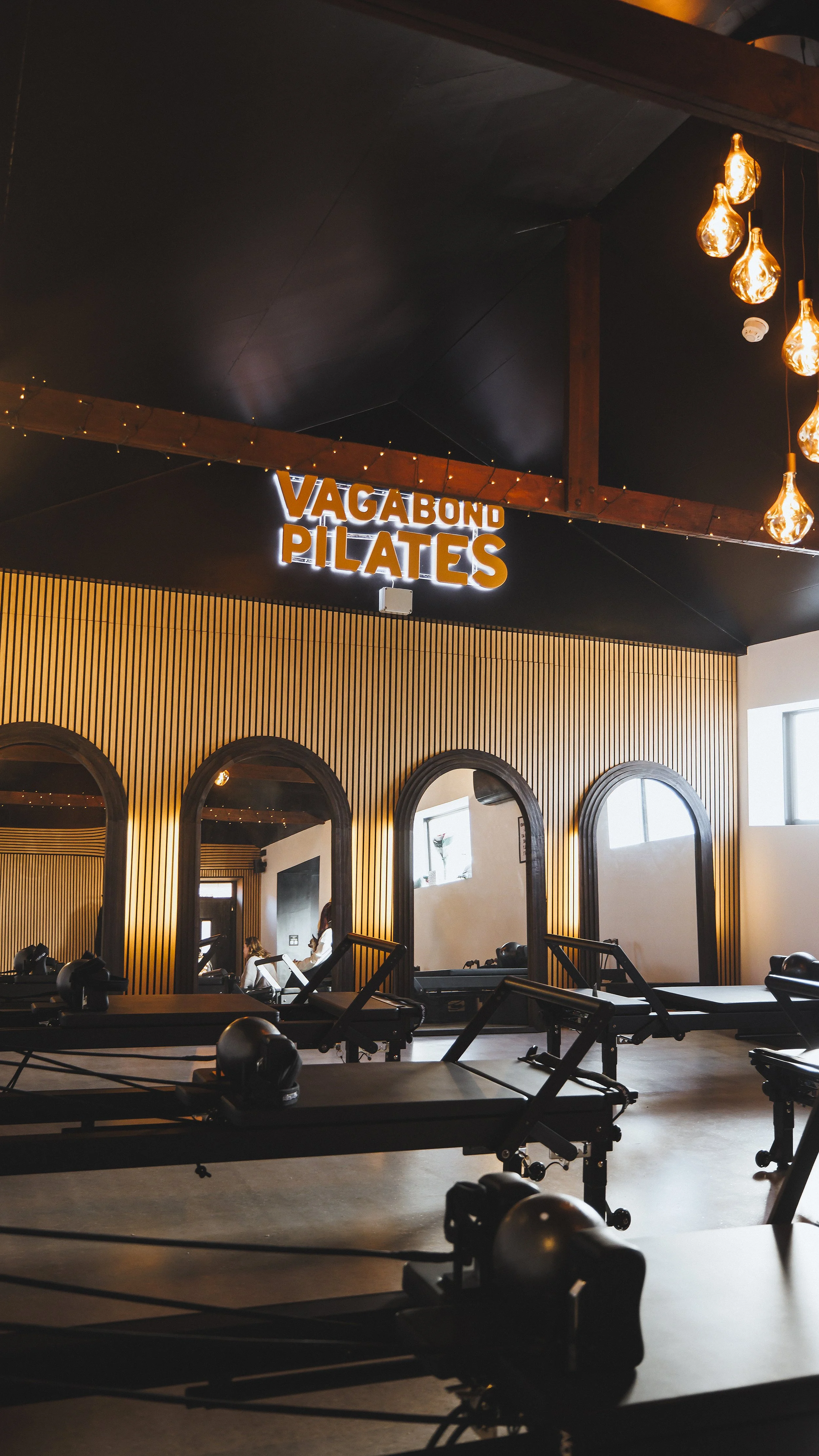 Interior of a fitness studio with Pilates reformers, wooden wall accents, arched mirrors, and a neon sign that reads 'Vagabond Pilates', illuminated warm lighting, and a few people practicing Pilates.