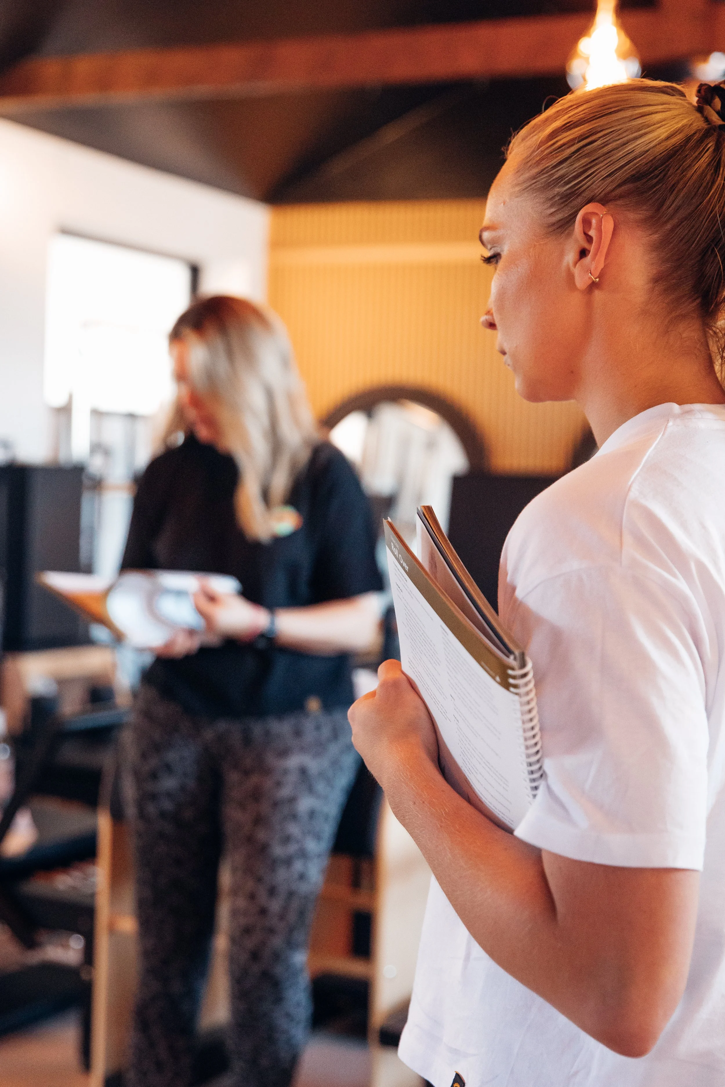 A woman with blonde hair tied in a ponytail, wearing a white t-shirt, holding a menu, looking at it, in a restaurant or cafe with warm lighting. Other people in the background are also present, blurred.