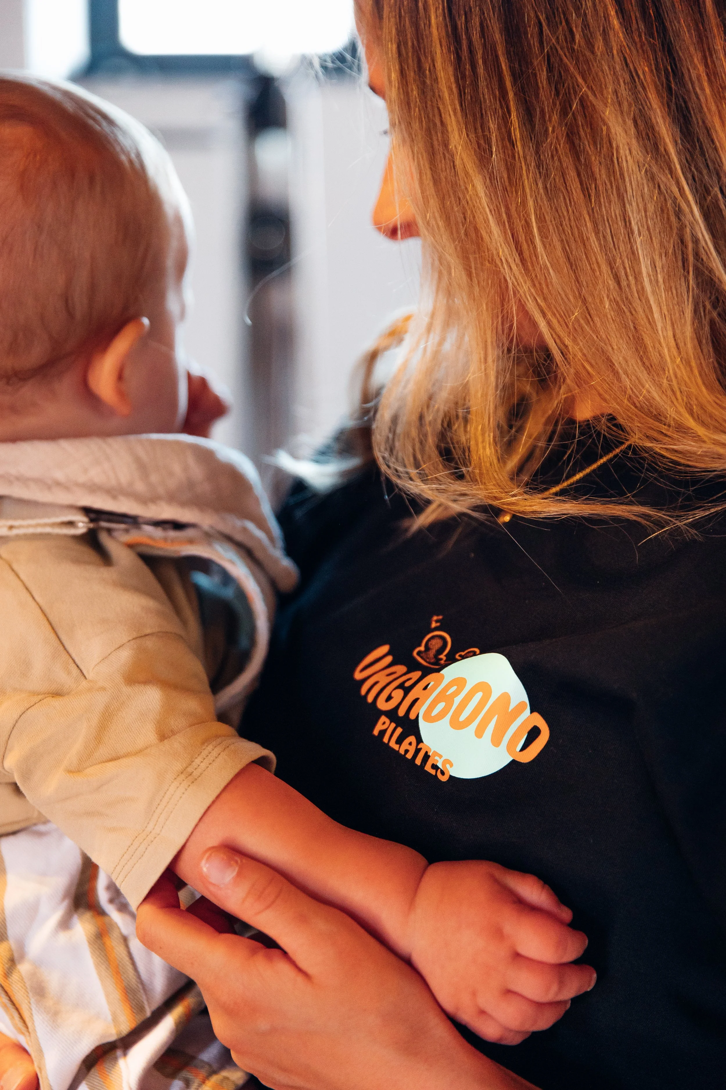 A woman with light brown hair holding a small child in her arms. The woman is wearing a black T-shirt with an orange and white logo that says "Canguro" and has a heart graphic. The child is wearing a beige jacket or hoodie, and they are close together, sharing a moment of affection.