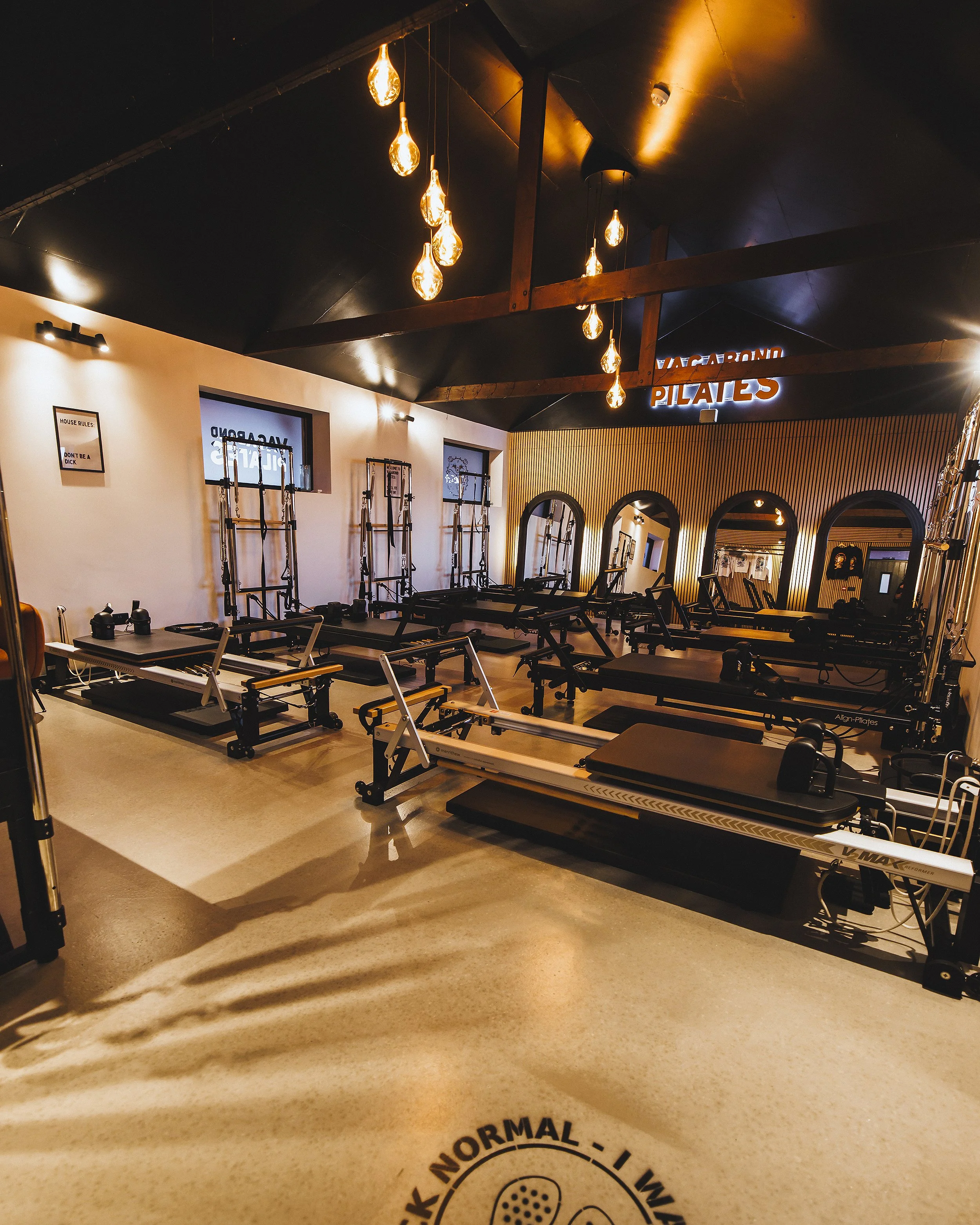 Fitness studio with Pilates reformer machines, hanging Edison bulb lights, mirror, wood panel wall, and a sign that says JACARAND PILATES.
