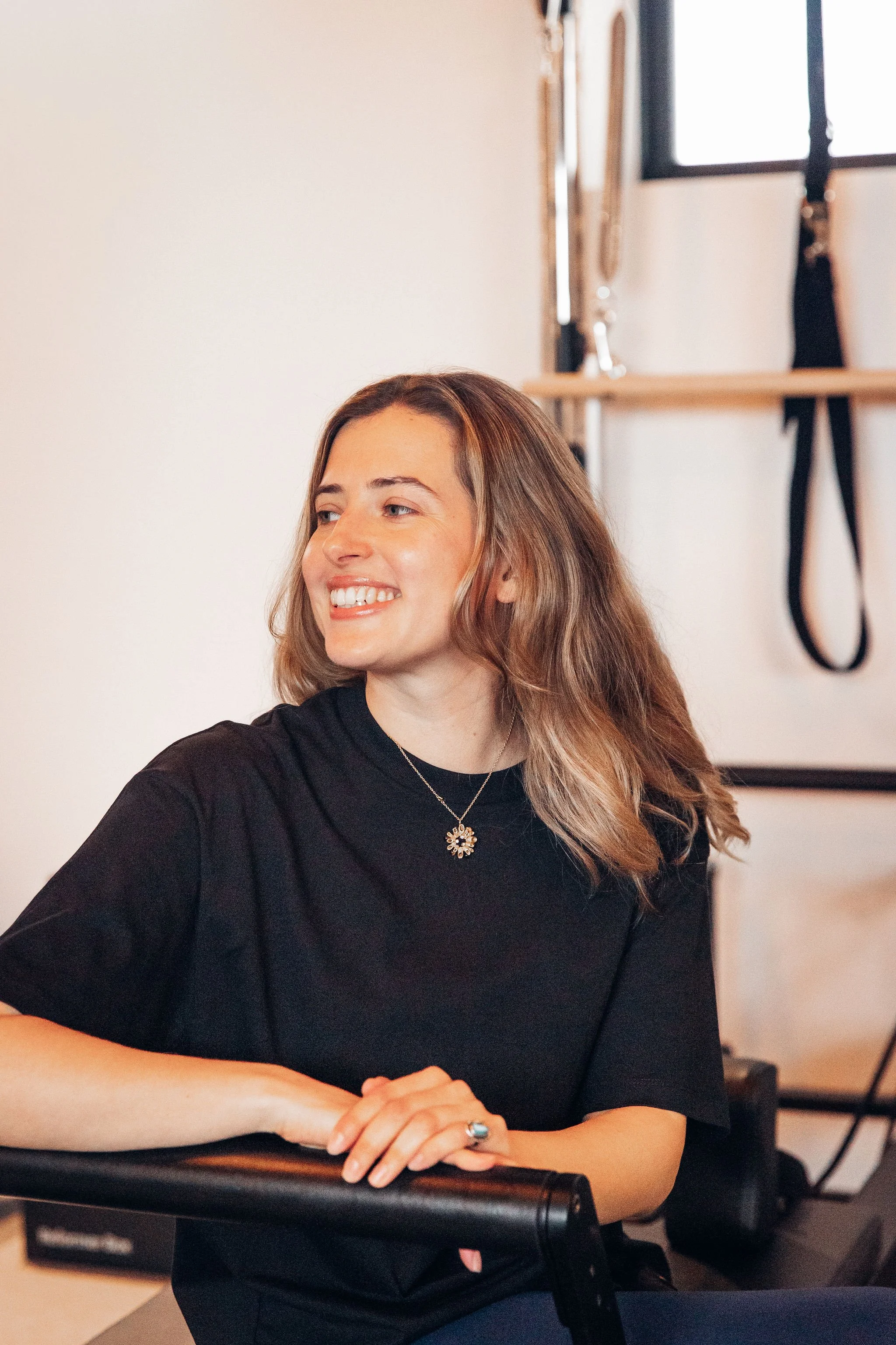 A woman with long, wavy, light brown hair smiling while sitting at a table in a room that has a window and gymnastic equipment in the background.