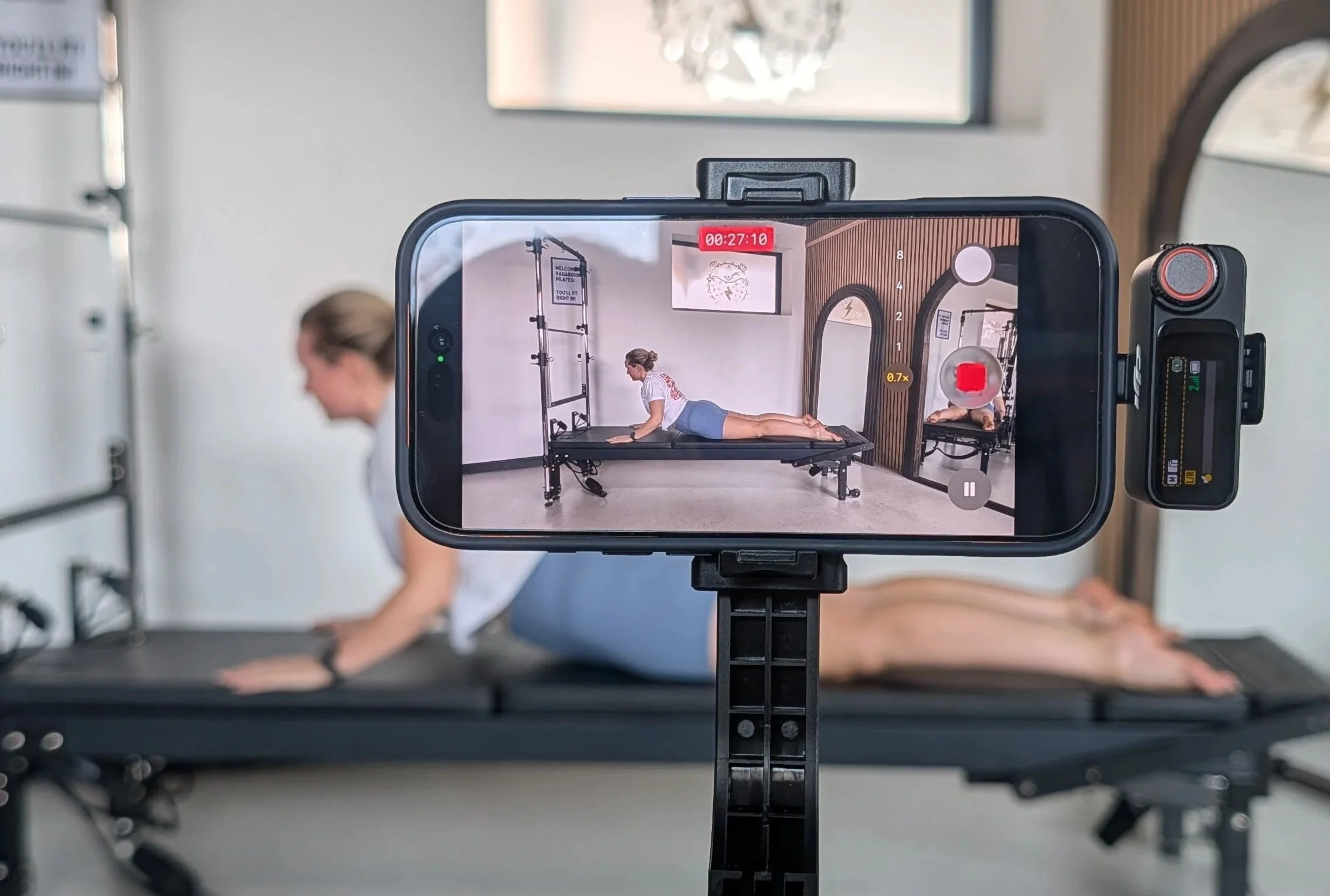 A woman performing a yoga pose on a yoga mat in a room with wooden wall panels and hanging decor, as seen through a smartphone camera's screen.