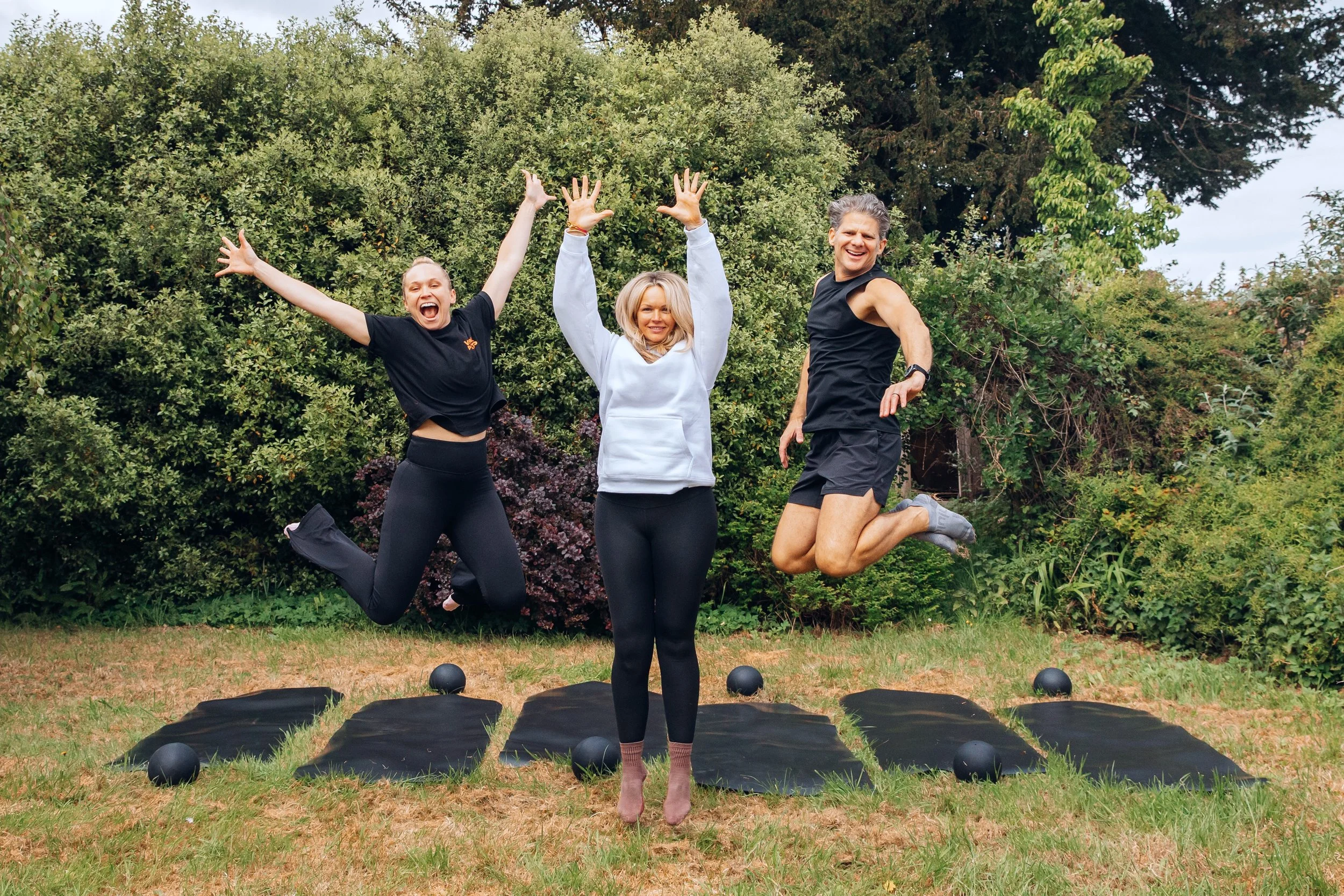 Three people jumping outdoors on a grassy area with trees in the background. They are smiling and appear to be exercising or celebrating. The person in the middle is a woman wearing a white hoodie, while the other two are a woman and a man wearing black. Exercise mats and black balls are on the ground.