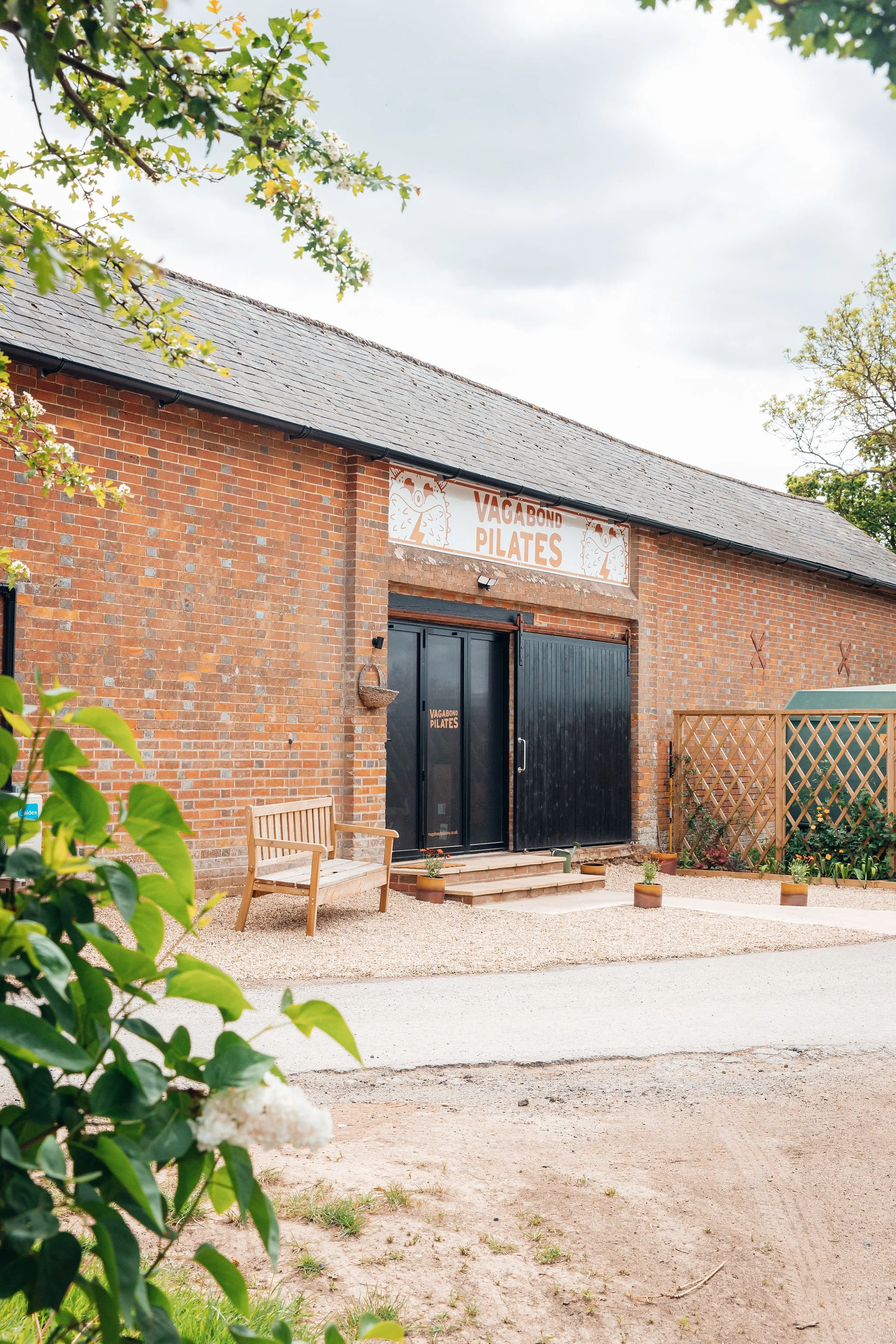 A brick building with a sign that says 'Vagabond Pilates' and an owl illustration. There's a black door, a small wooden bench, potted plants, and a wooden fence. The foreground has green leaves and white flowers. The ground is gravel or dirt, and the sky is cloudy.