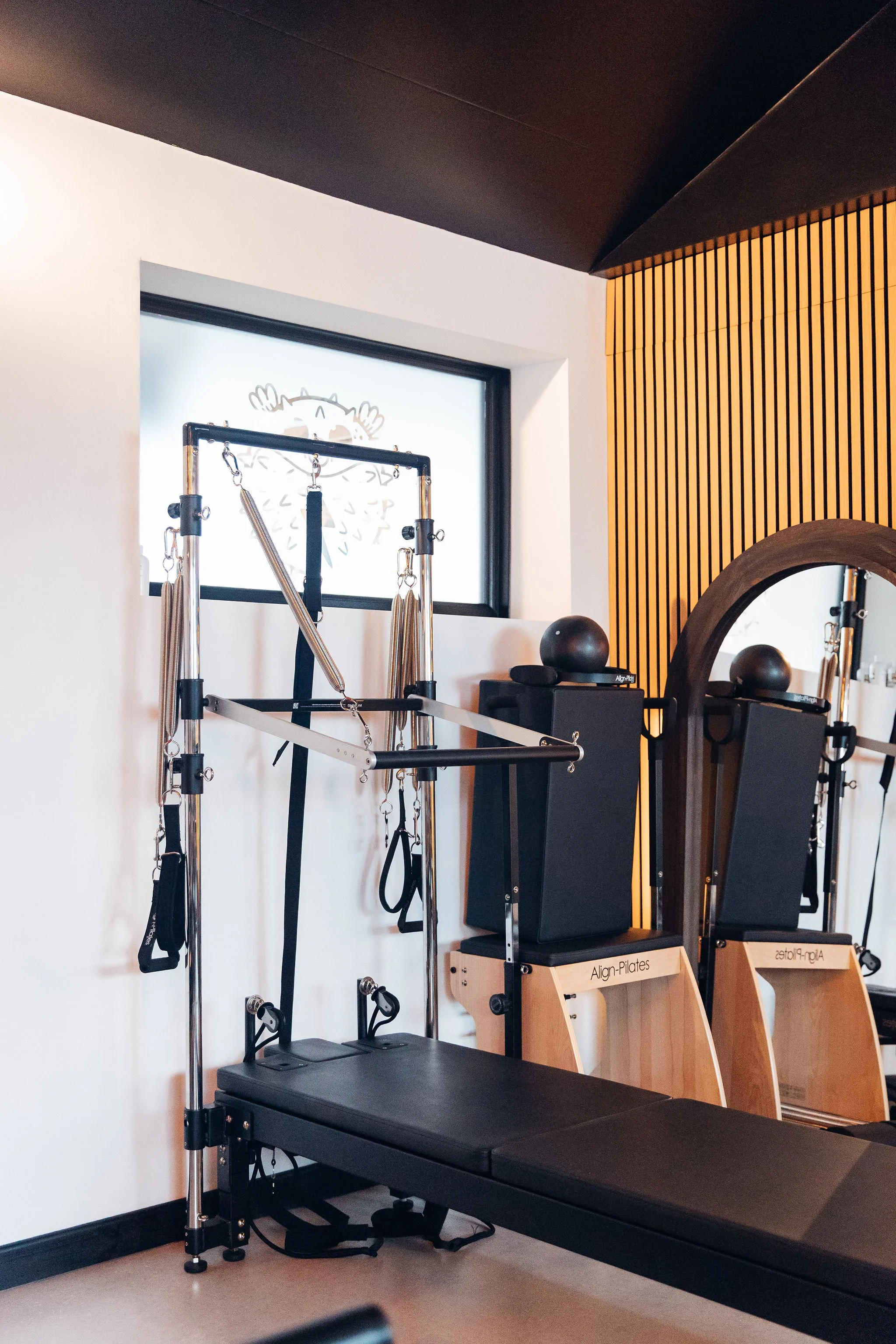 Pilates reformer machine, Pilates chair, and wooden Pilates boxes in a well-lit Pilates studio with wooden wall panels and a window with a cartoon reindeer drawing.