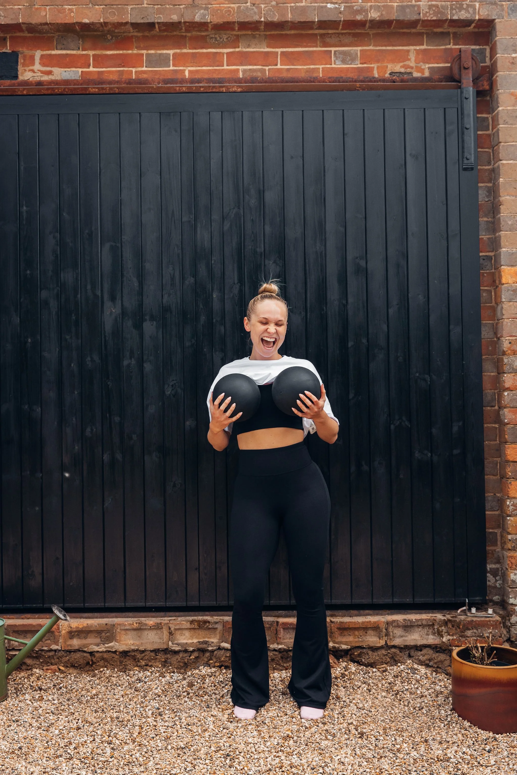 A woman with a top knot hairstyle, wearing black leggings and a white crop top, is smiling and holding two black exercise balls in front of a black wooden door and brick wall background.