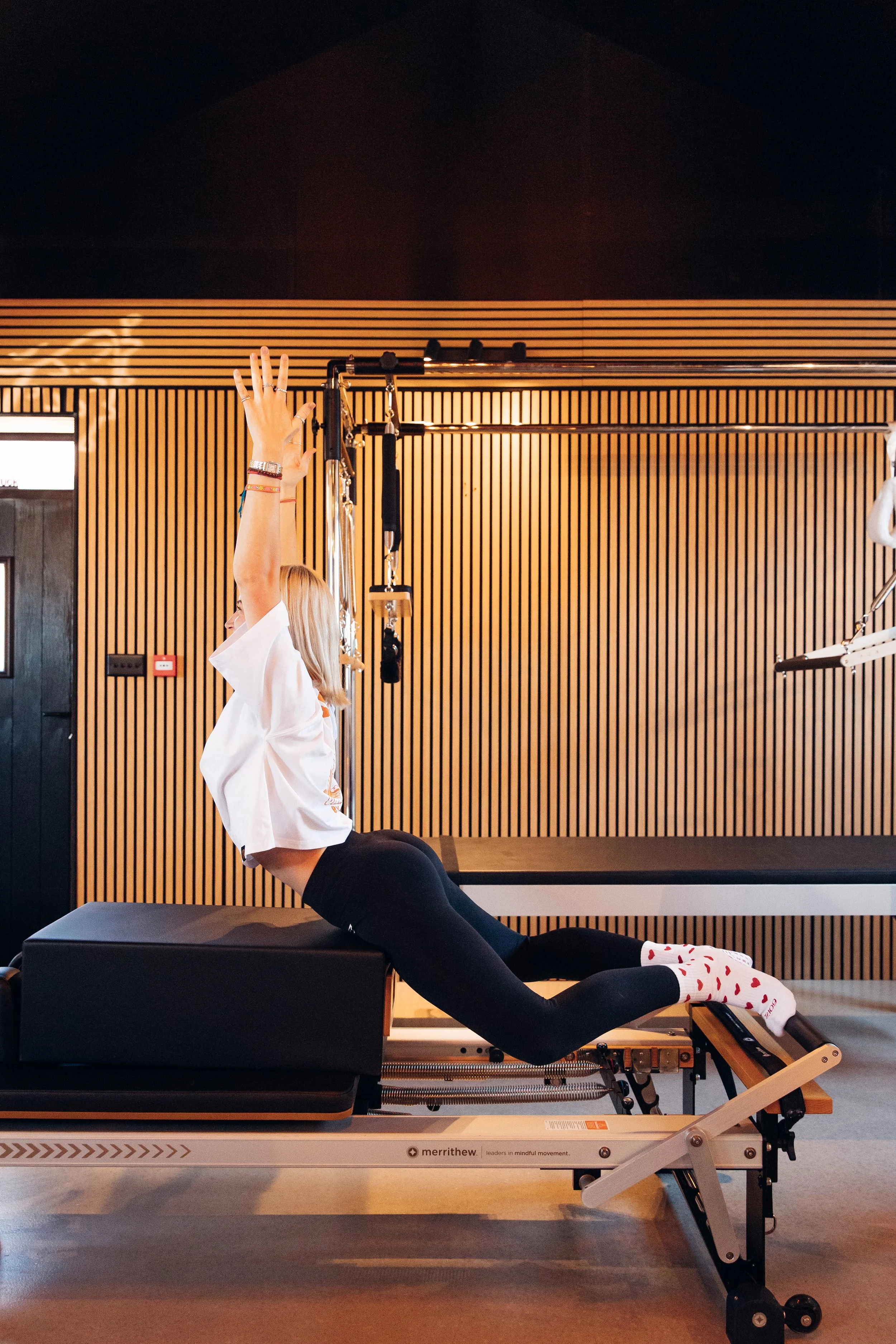 A woman in a white shirt and black leggings performs a yoga pose on a medical or therapy table in a room with wooden wall panels. She is in a front-leaning lunge position with one knee on the table and the other leg extended back. She raises her arms above her head, reaching upwards, and wears colorful bracelets and multiple rings. The room appears to be a therapy or fitness space.
