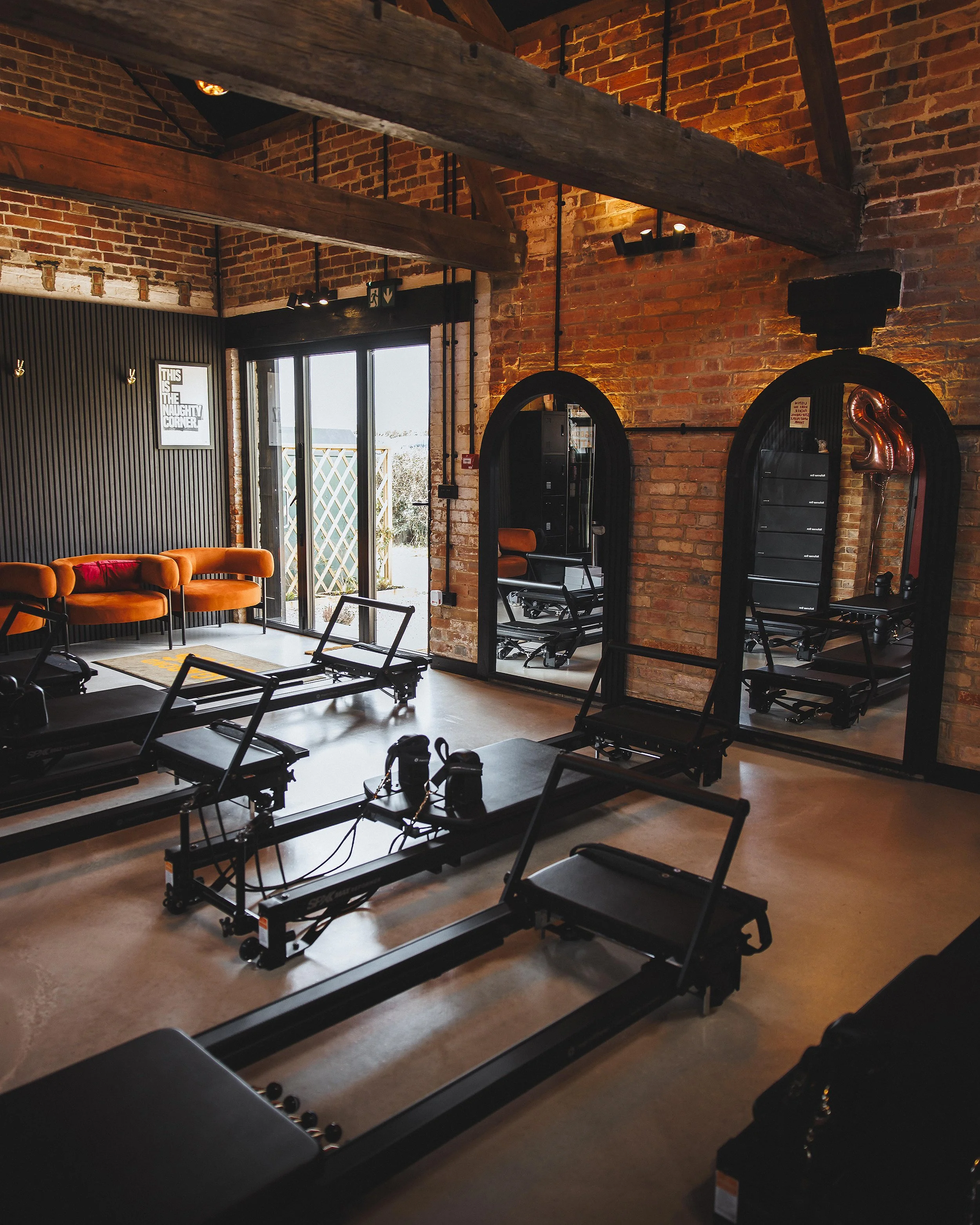 Indoor fitness studio with wood beam ceiling, brick walls, pilates reformer machines, orange cushioned chairs, and a glass door leading to an outdoor area.