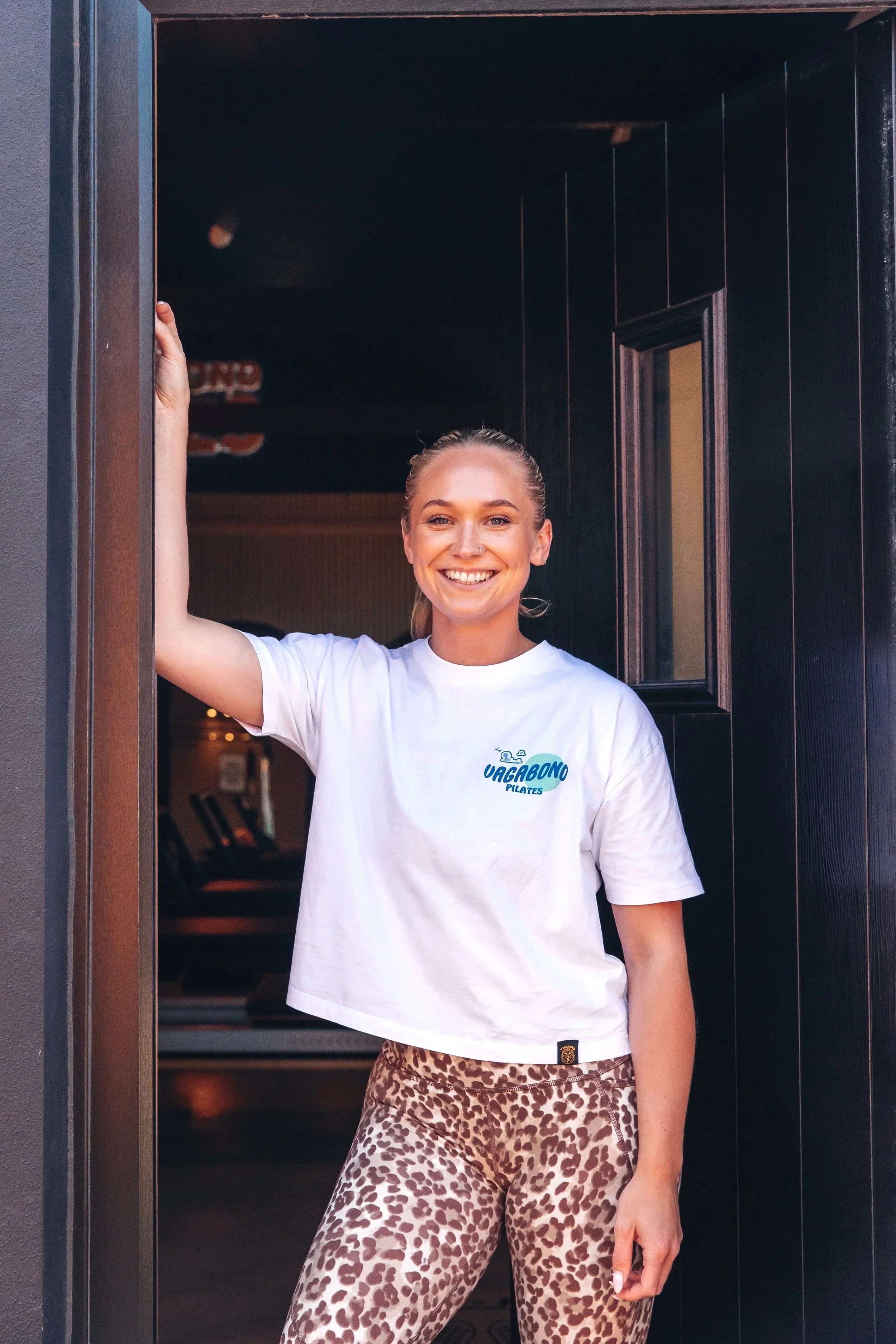 A smiling woman with blonde hair, wearing a white t-shirt with a logo, leopard print pants, standing in an open doorway.