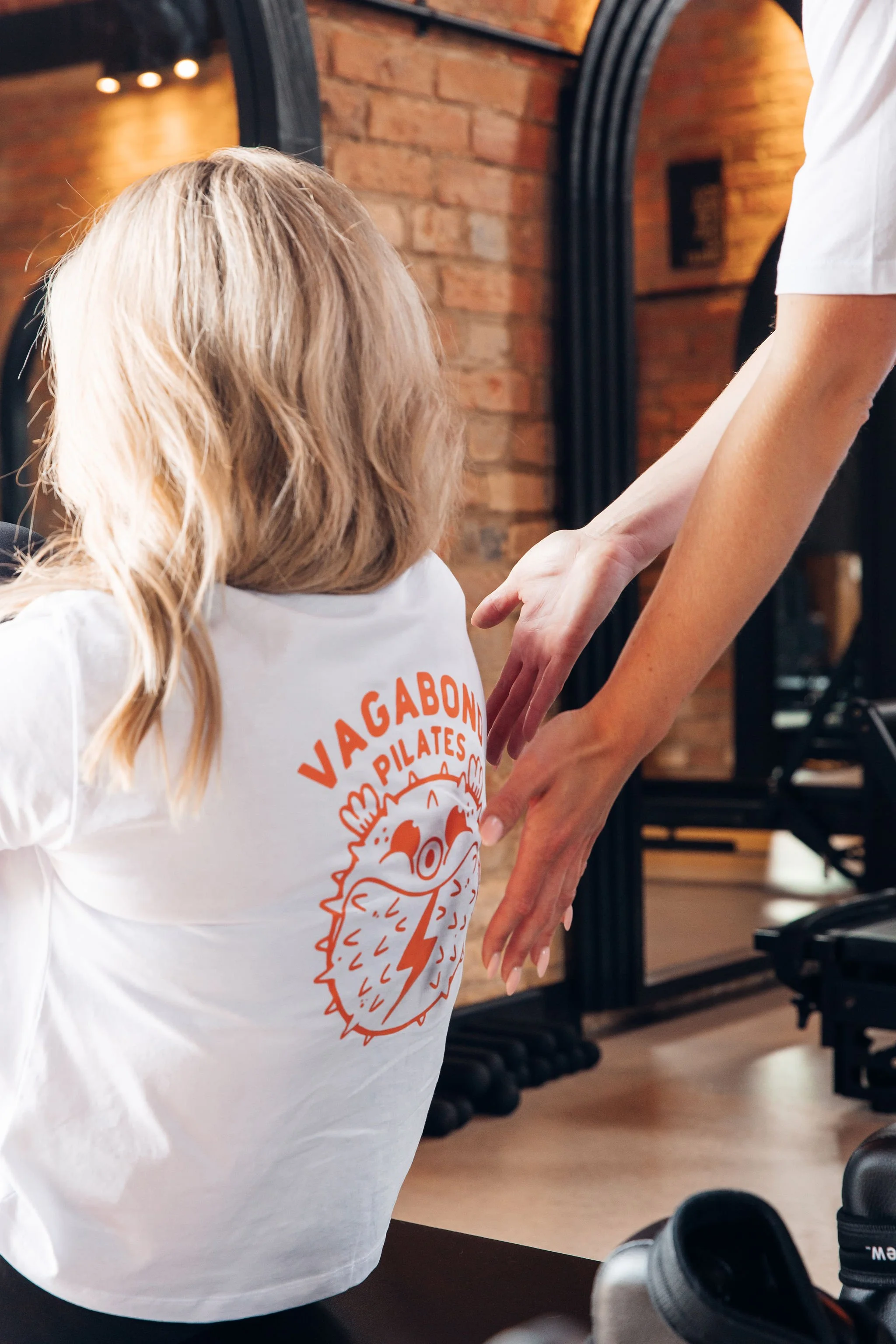 A person with blonde hair wearing a white T-shirt with an orange graphic of a dinosaur and the words "Vagabond Pilates" on it, holding or guiding a child's arm during a Pilates class in a gym with brick walls and gym equipment in the background.
