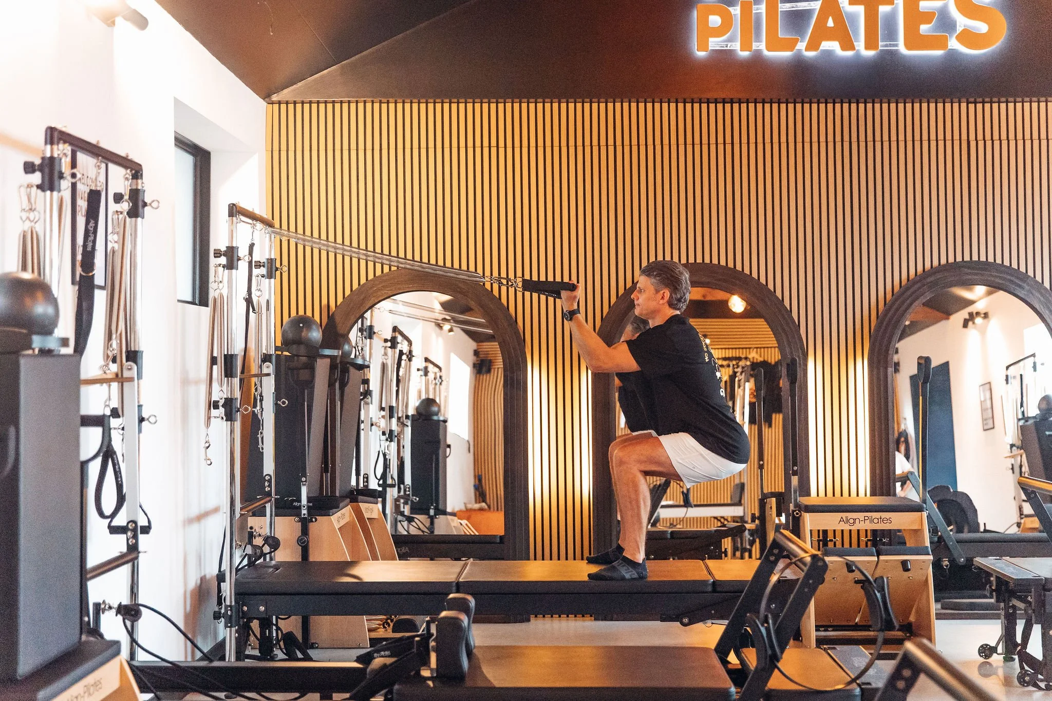 A man performing a squat pose on a Pilates reformer machine in a fitness studio, with wooden wall panels and a sign that says 'PILATES' illuminated in the background. Exercise equipment is visible in the room.