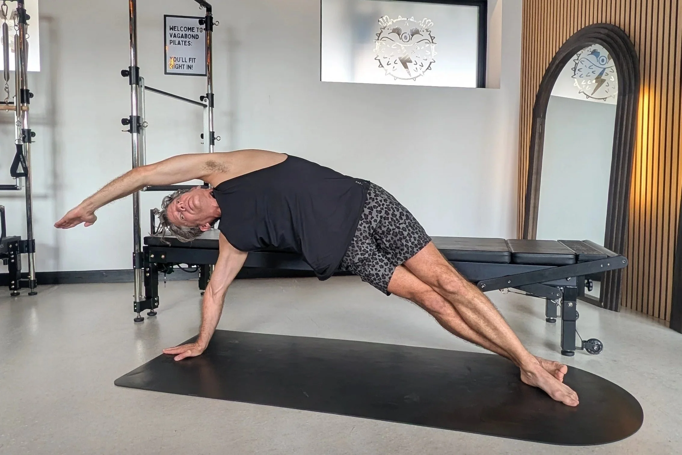 A man in a black tank top and leopard print shorts performing a yoga stretch on a black mat in a fitness studio with a heated pole, a large mirror, and cartoonish artwork of a creature with a lightning bolt on its face on the wall.