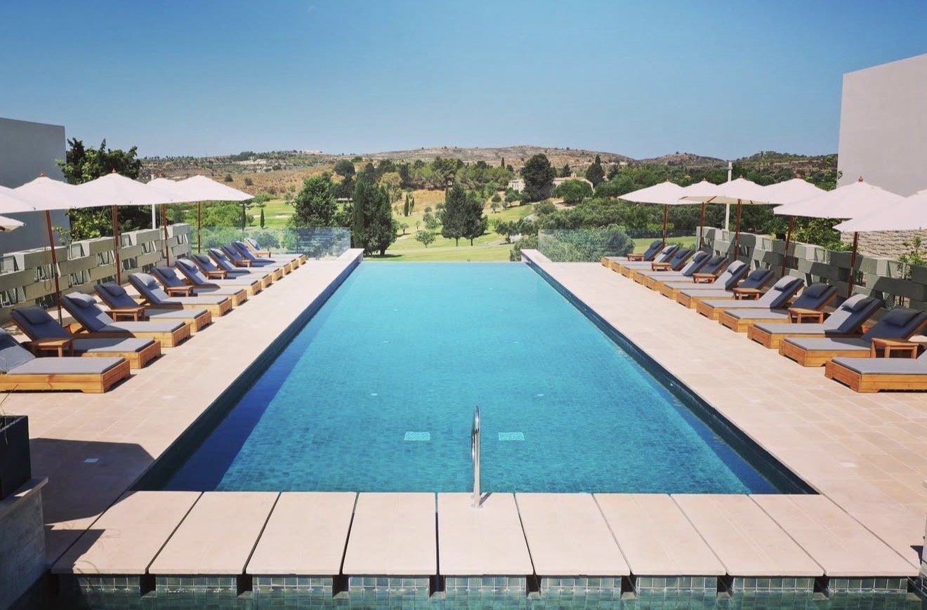 Outdoor swimming pool with lounge chairs and umbrellas on either side, overlooking a lush green landscape with trees and rolling hills under a clear blue sky.