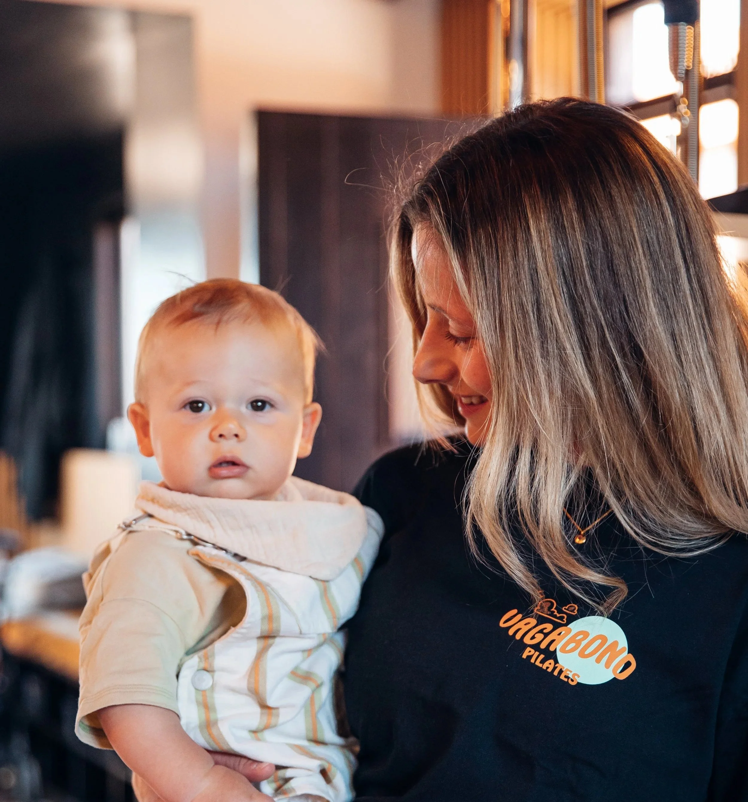 A young child with red hair and blue eyes looking at the camera, being held by an adult with long, straight, dark blonde hair, indoors near a window with sunlight. The adult's black shirt has an orange logo that says "Vagabond Pilates".