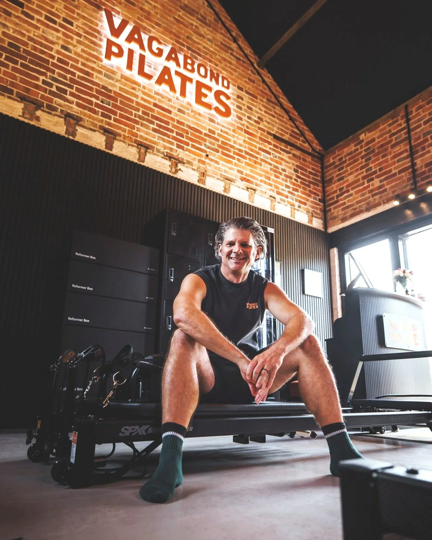 A man in workout clothes sitting on a foam roller in a gym with an industrial brick wall. Behind him is a sign that reads "Vagabond Plates" and exercise equipment.