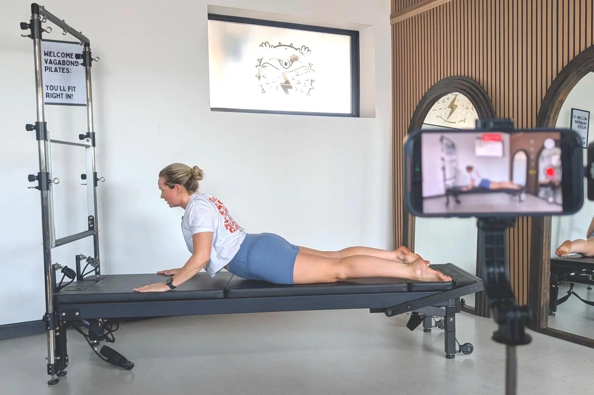 A woman performing a plank exercise on a Pilates reformer machine in a fitness studio, with a man filming her using a camera on a tripod.