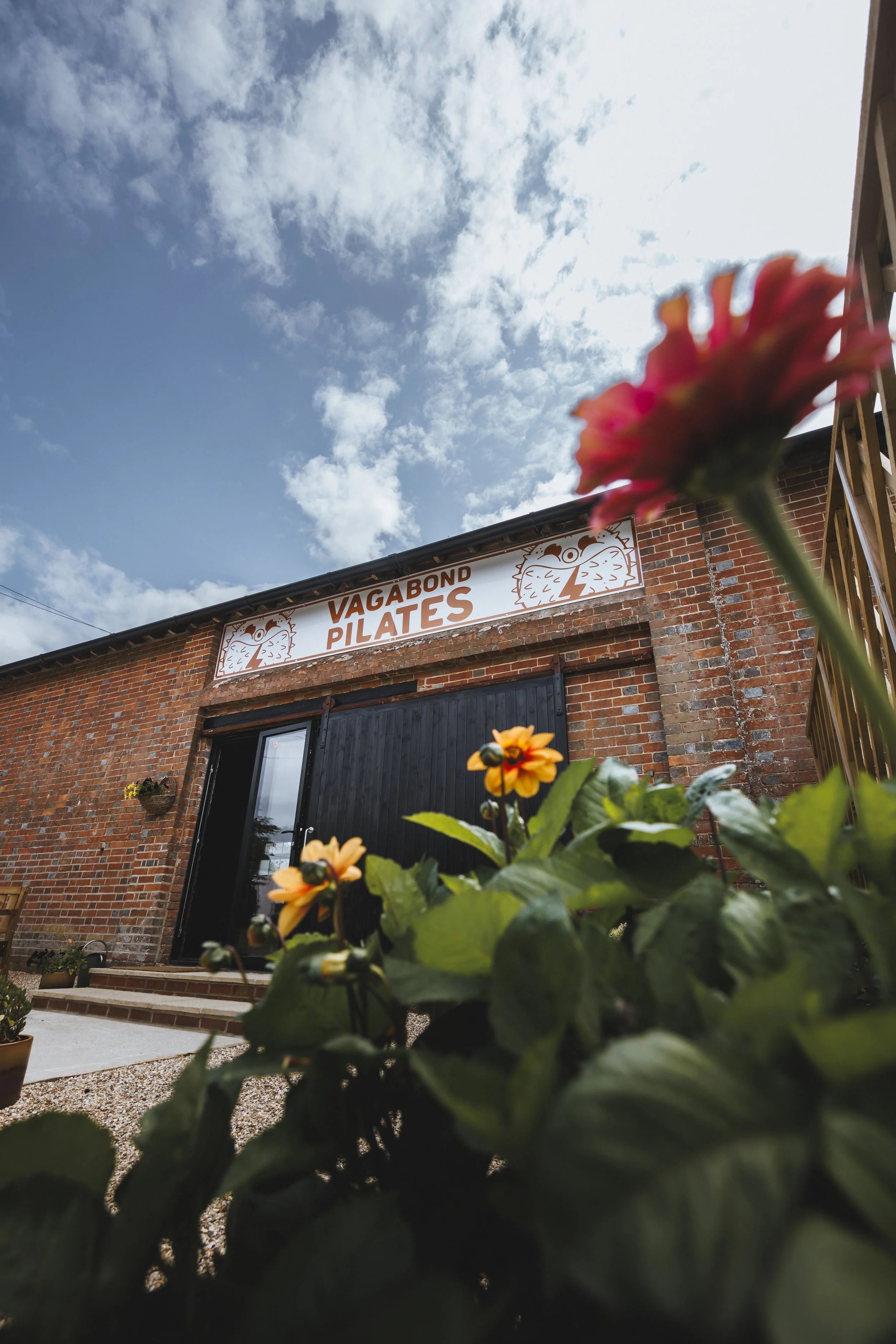 A brick building with a sign that reads "Vagabond Pilates" in front of a partly cloudy sky, with some flowers in the foreground.