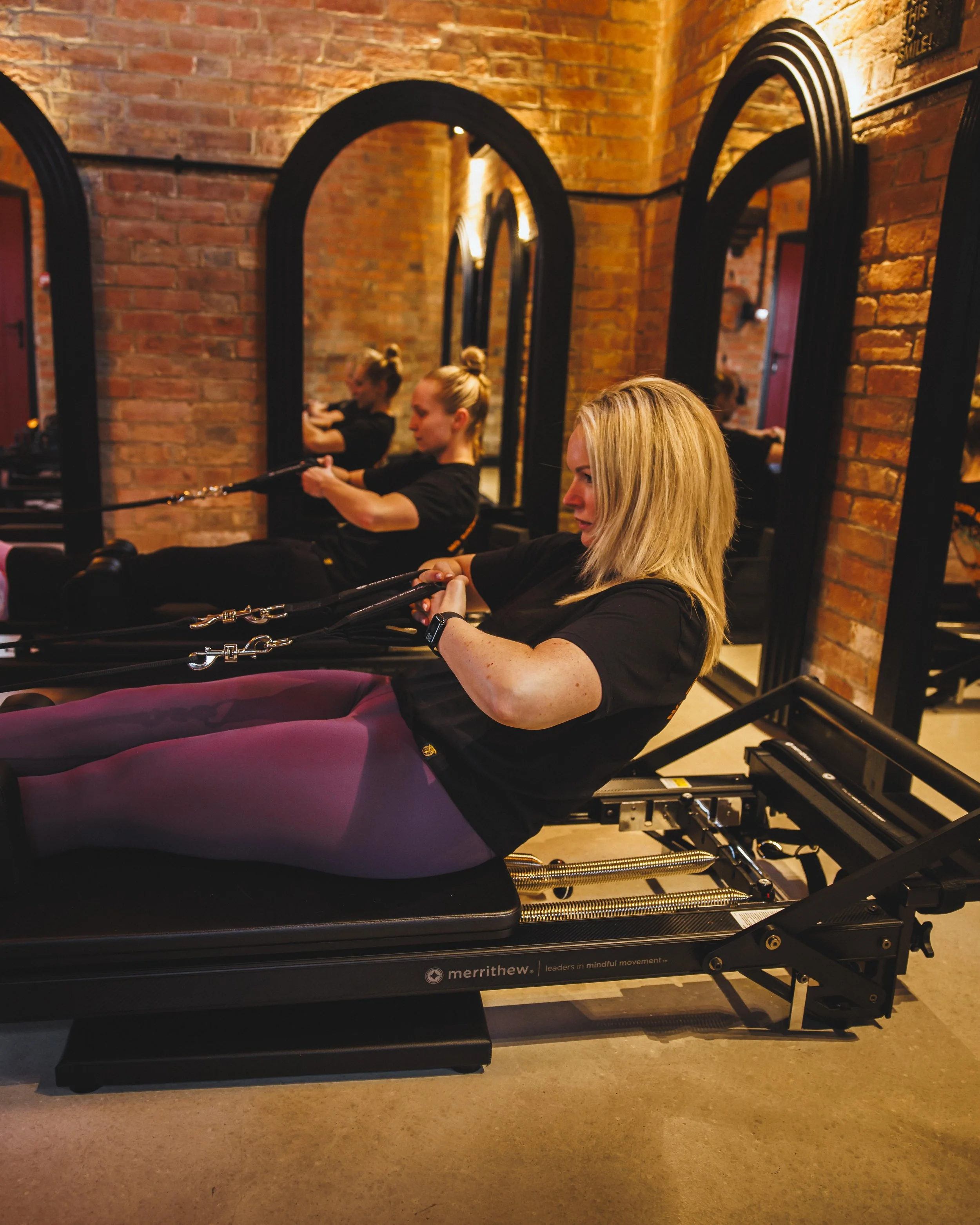 Two women exercising on a Pilates reformer machine in a brick-walled fitness studio. One woman has blonde hair and is sitting on the reformer, holding onto the straps. The other woman has her hair tied up and is also using the reformer, focusing on her exercise. The room has warm lighting and mirrors reflected on the wall.