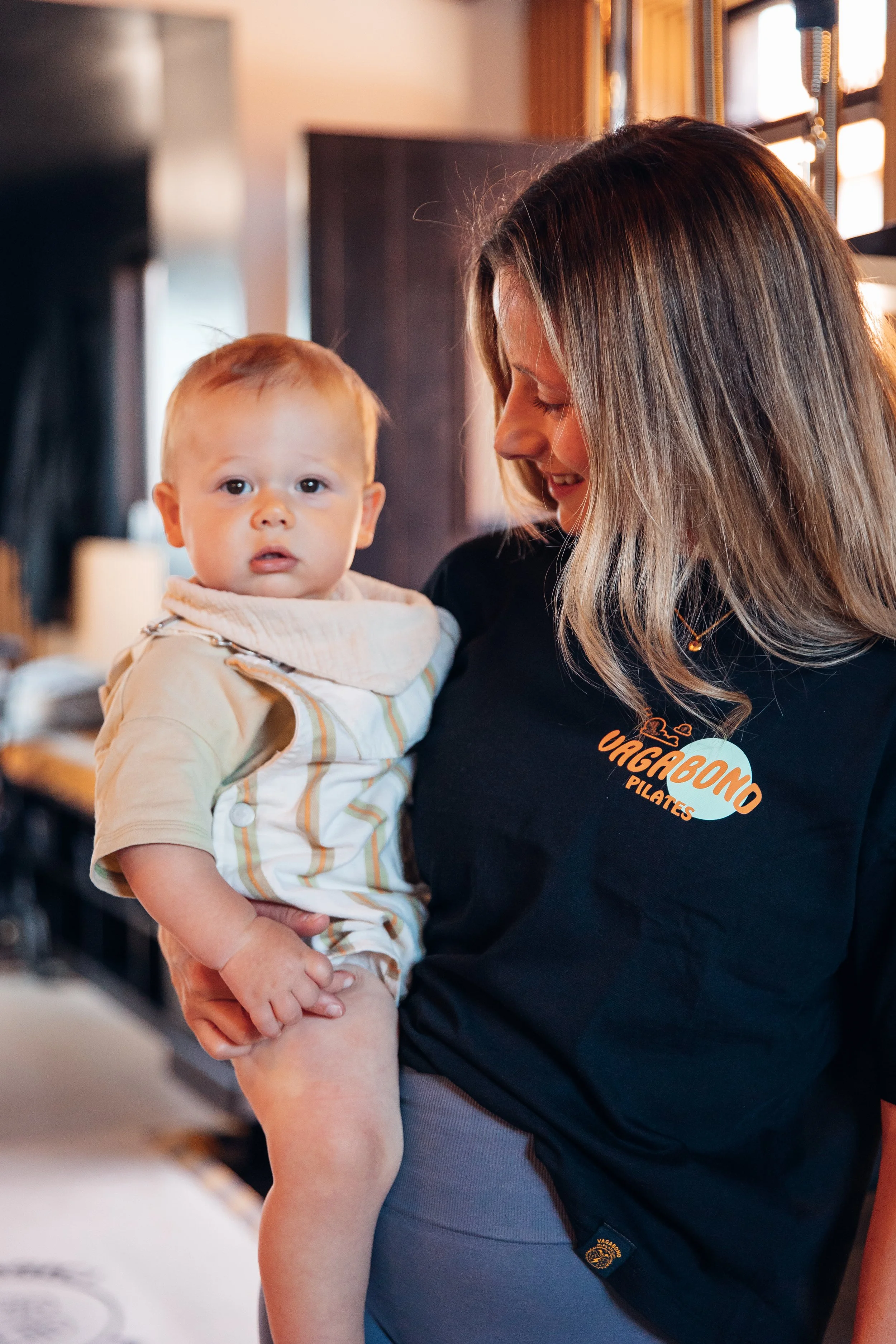 A smiling woman with long brown hair holding a small child with red hair in a pilates fitness studio. The woman is wearing a black shirt with a sticker that says "VAGABOND PILATES." The child is wearing a light-colored hoodie with light stripes.