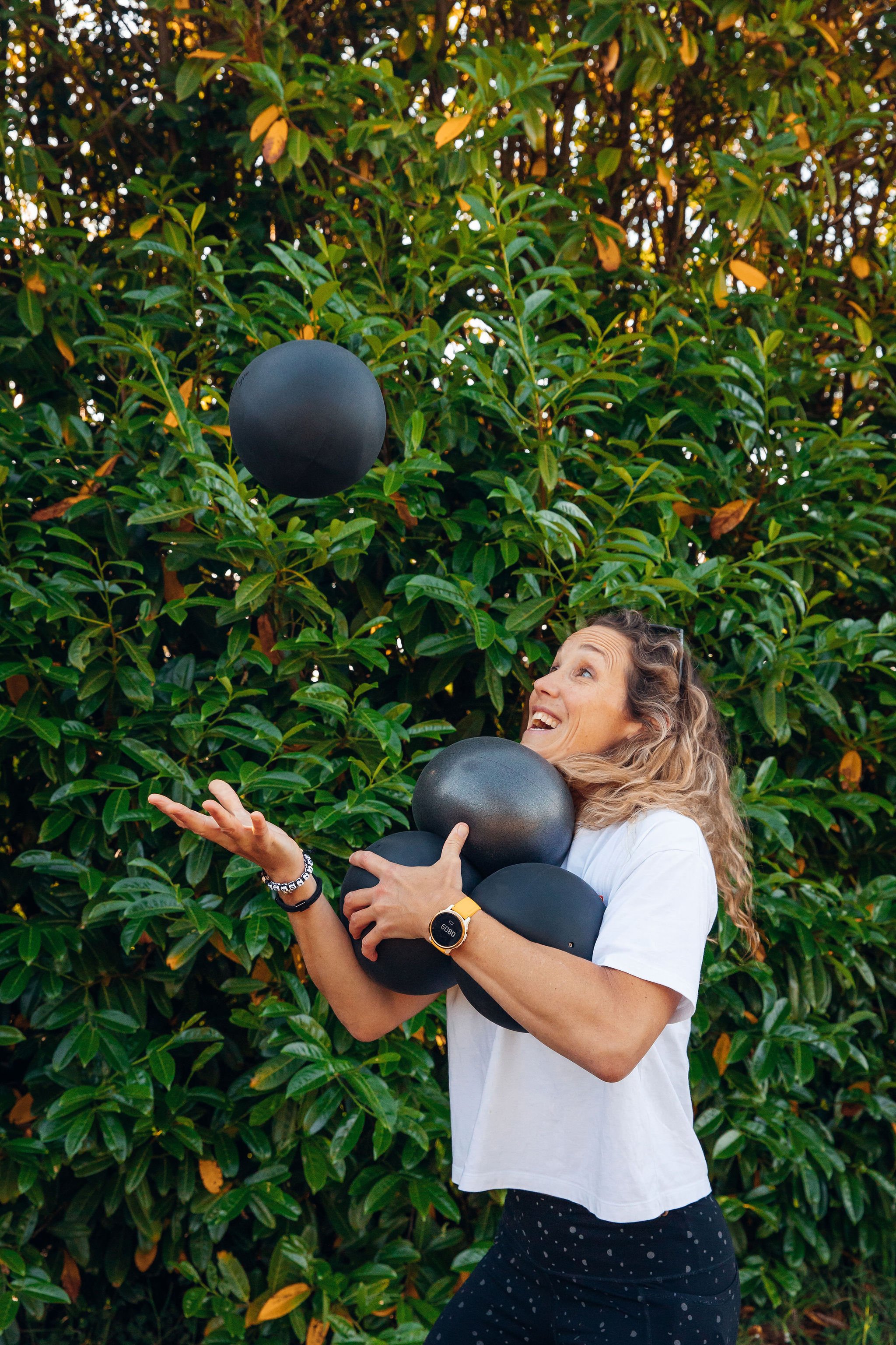 A woman with long curly hair wearing a white t-shirt and black polka dot pants, holding black balloons, standing in front of green leafy bushes, smiling and looking up.