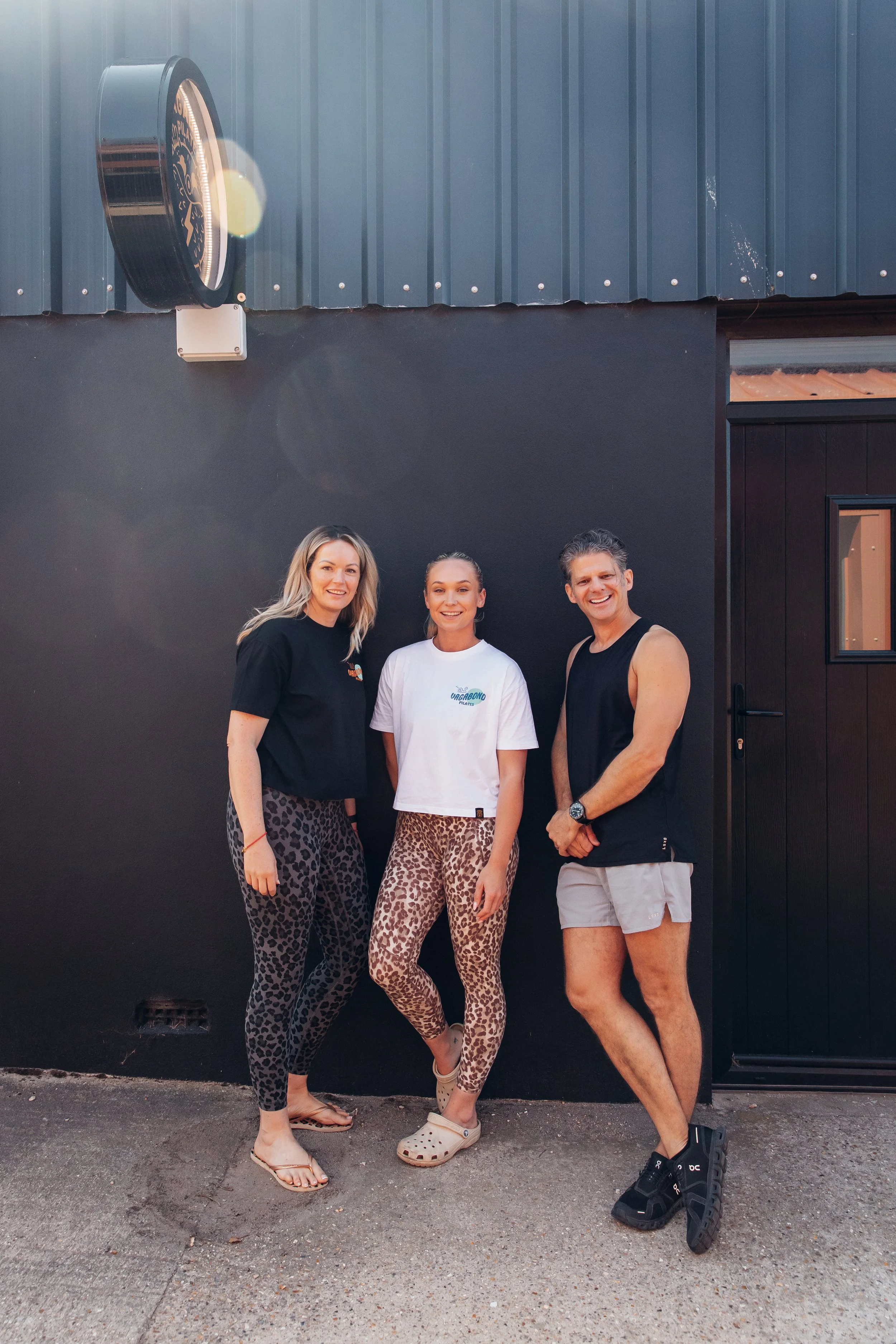 Three people standing together outside in front of a black wall with a large building or window to the right. The two women are wearing petal leggings in animal print, one in black and gray and the other in brown and beige. The man is dressed in a sleeveless black shirt and light-colored shorts, all smiling at the camera.