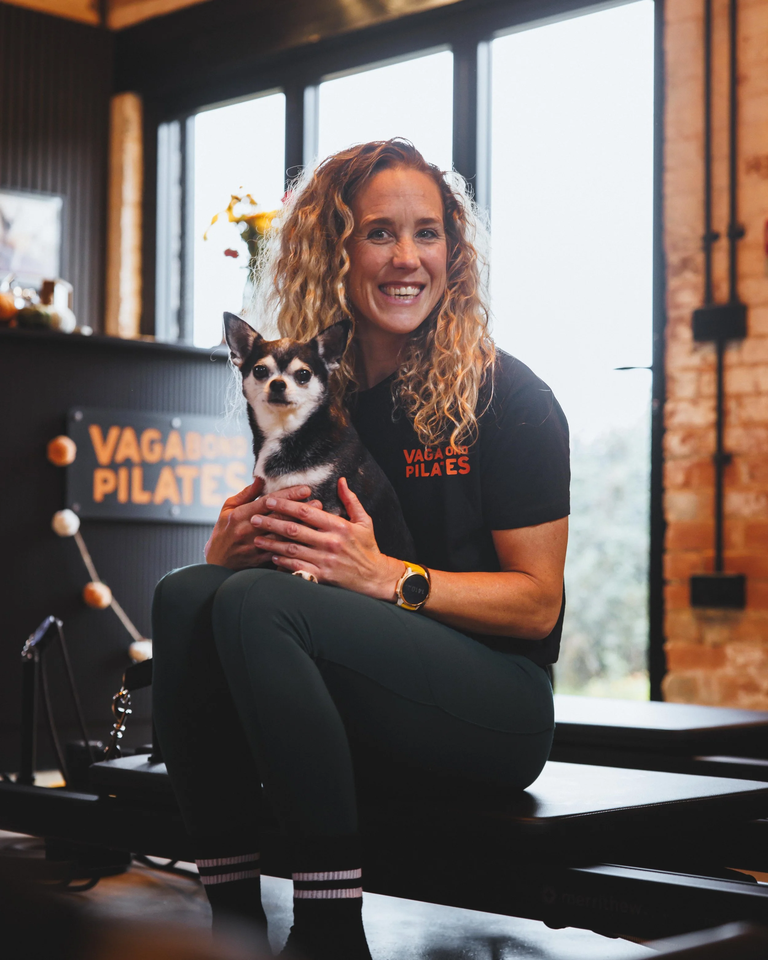 A woman with curly red hair sitting on a Pilates reformer machine holding a small black and white dog, in a Pilates studio with large windows and brick walls.