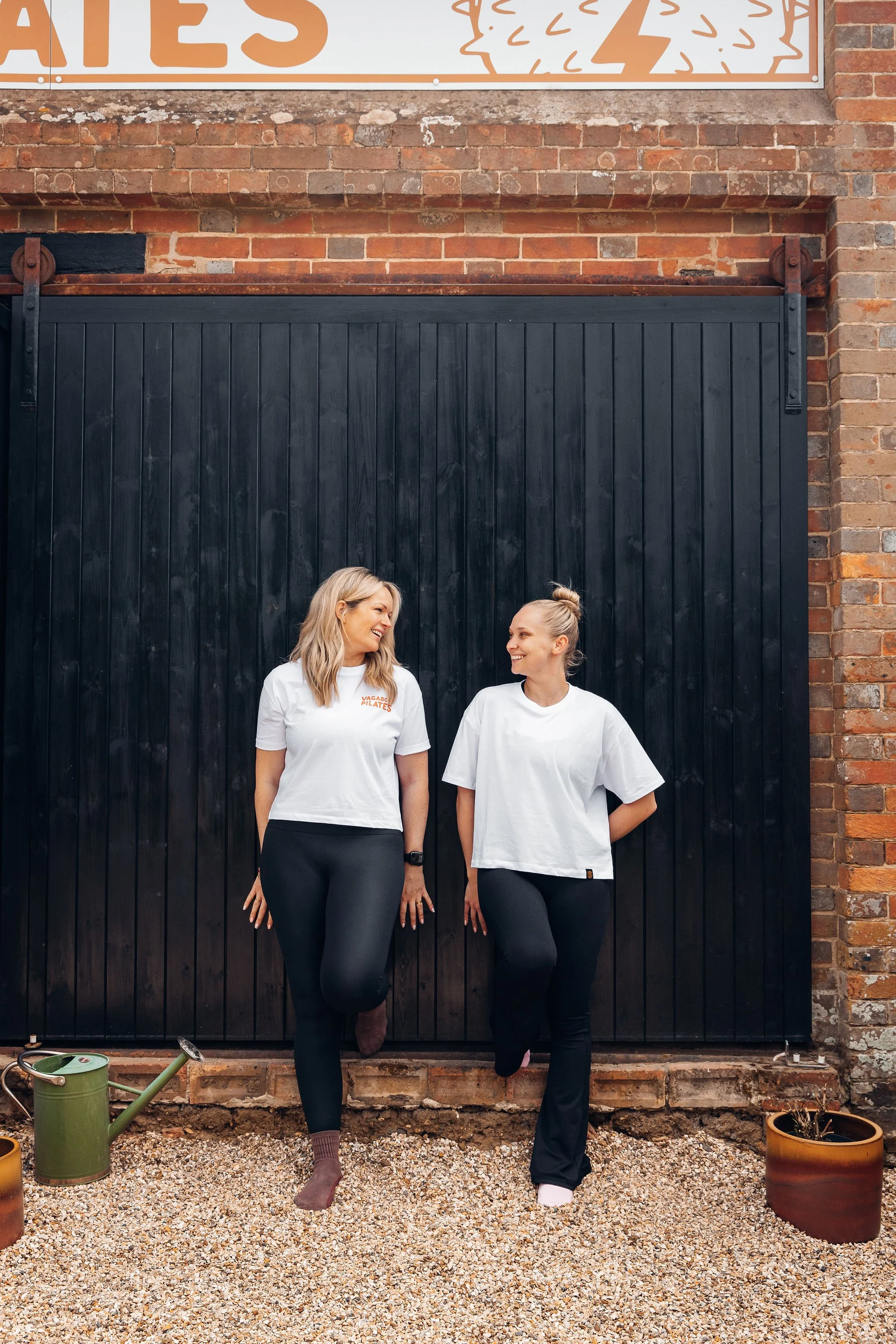 Two women standing in front of brick wall with a black sliding door, smiling and looking at each other. One woman has blonde hair, wearing a white t-shirt with orange text and black pants. The other woman has hair tied up in a bun, wearing a white t-shirt and black pants.