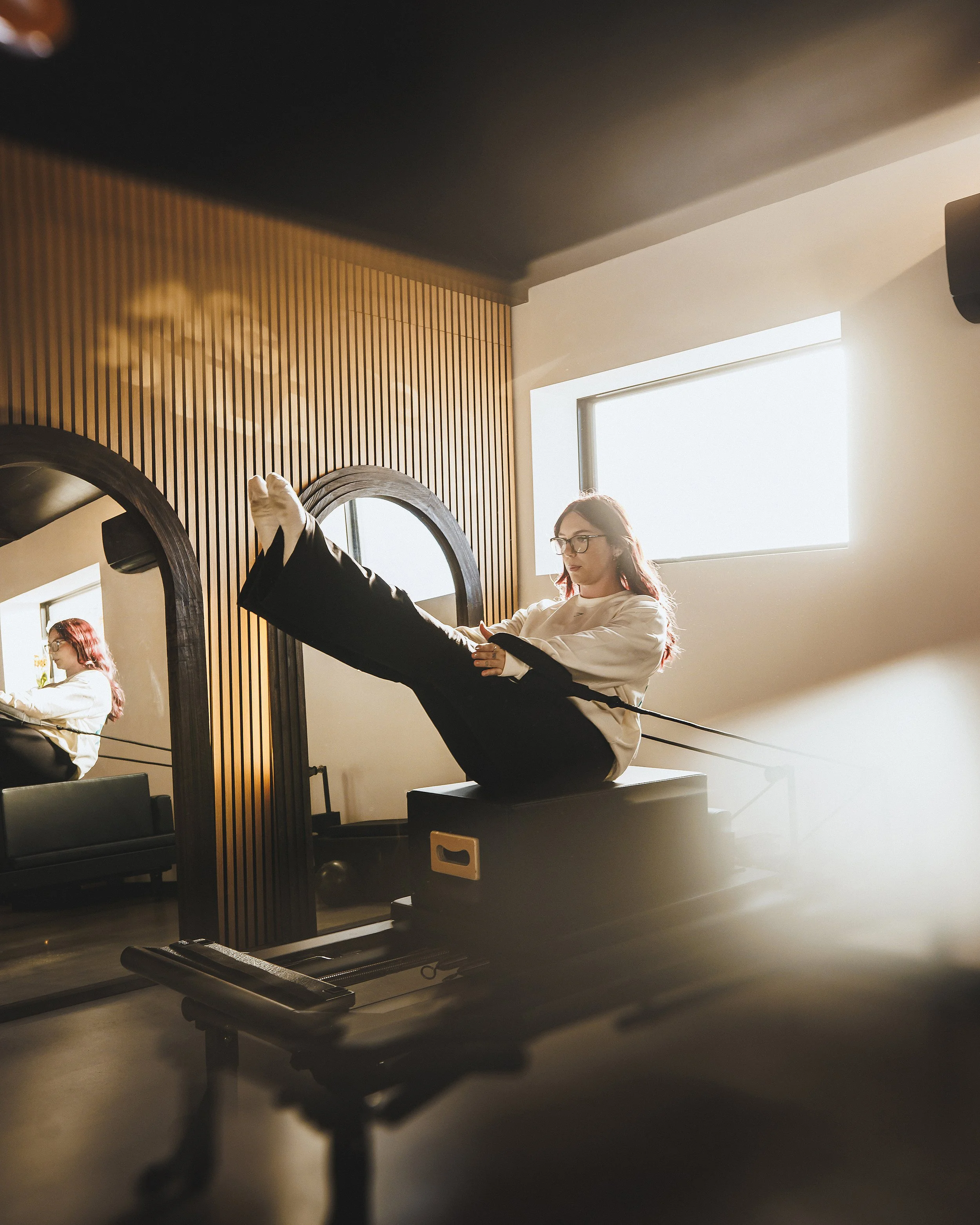 A woman is practicing yoga on a black mat inside a room with wooden wall paneling and a large mirror. She is sitting on a platform with one leg raised, holding her foot with one hand and her knee with the other. She is wearing a white long-sleeved shirt and black pants. The room has a window with sunlight streaming in.