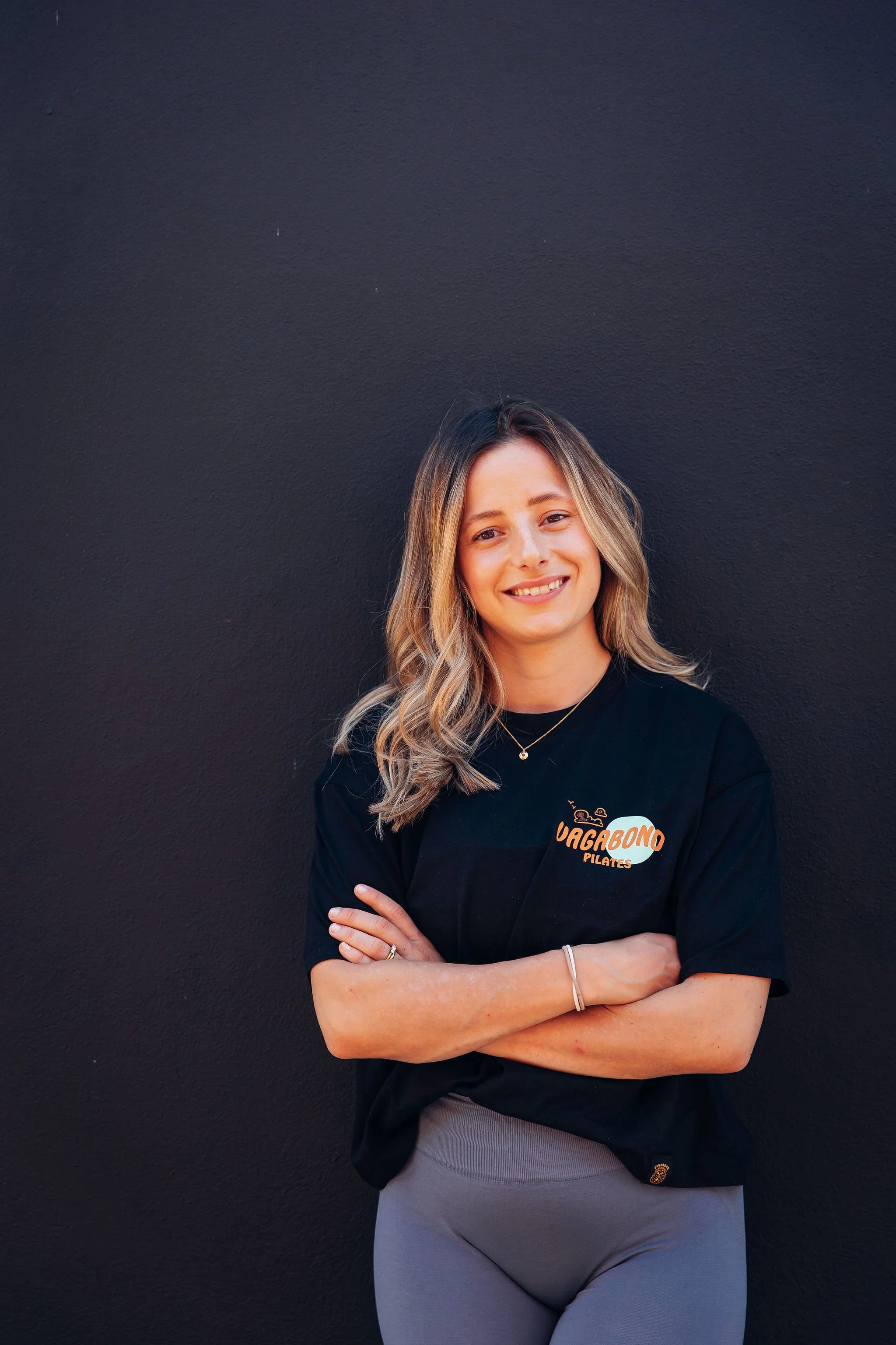 Young woman with long blonde hair standing against a black wall, smiling, wearing a black t-shirt with an orange and white logo that reads "VAGABONO PILATES," clasping her arms across her chest, and wearing gray leggings.