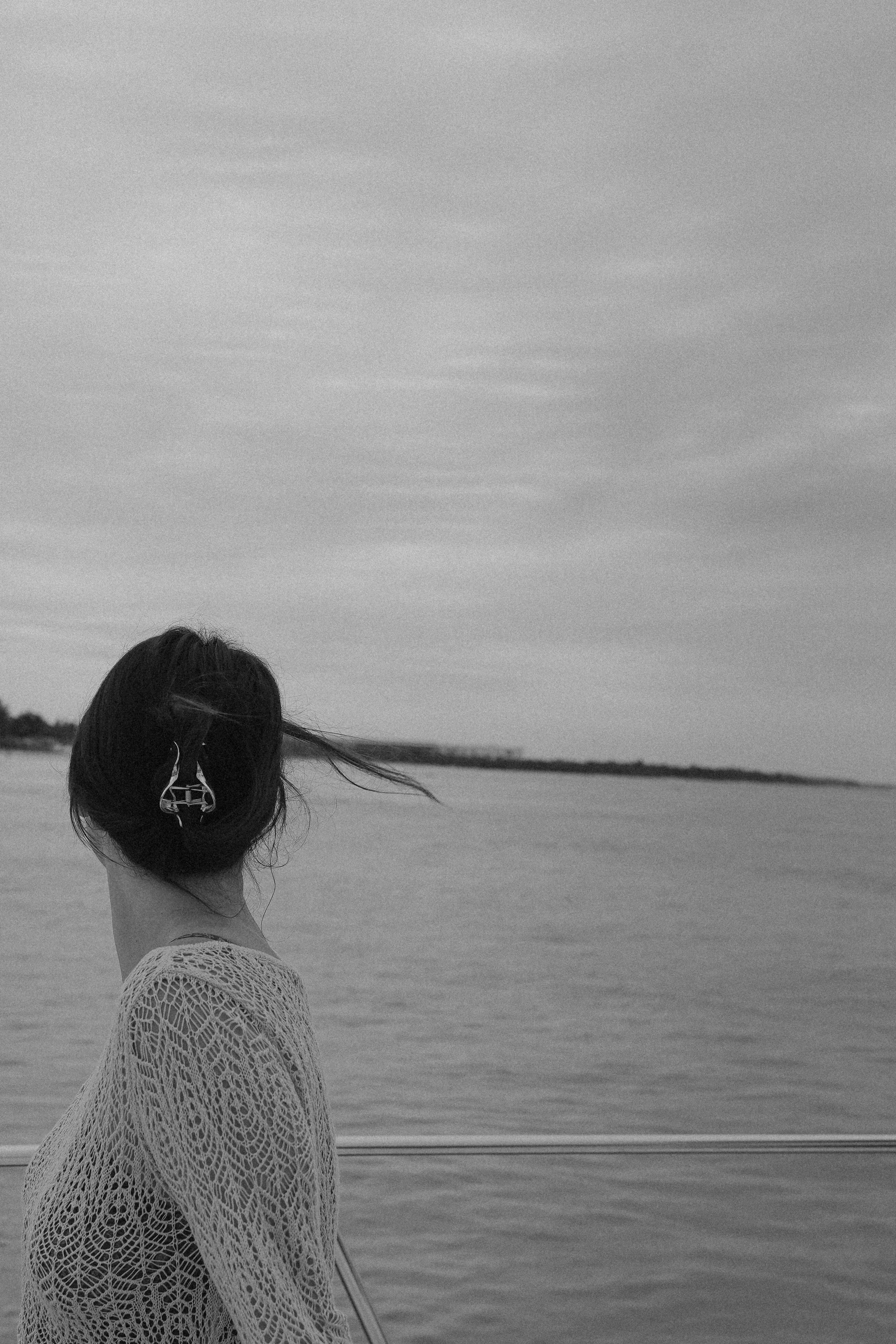 A woman with dark hair, wearing a crochet cover-up, standing on a boat or dock by a large body of water, looking away from the camera with her hair slightly blowing in the wind. The image is in black and white.