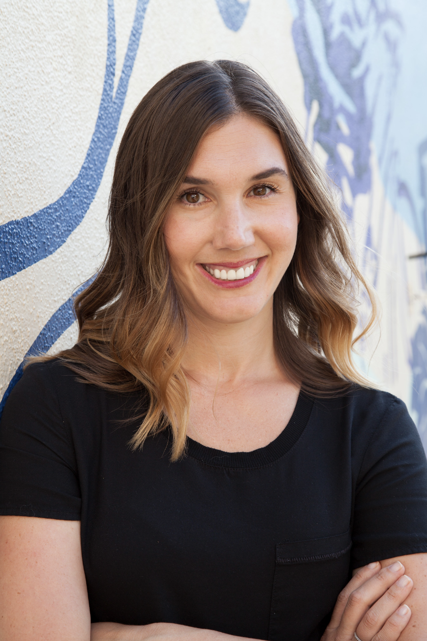 A woman with wavy, shoulder-length hair smiling and standing with her arms crossed in front of a textured wall with blue graffiti art.