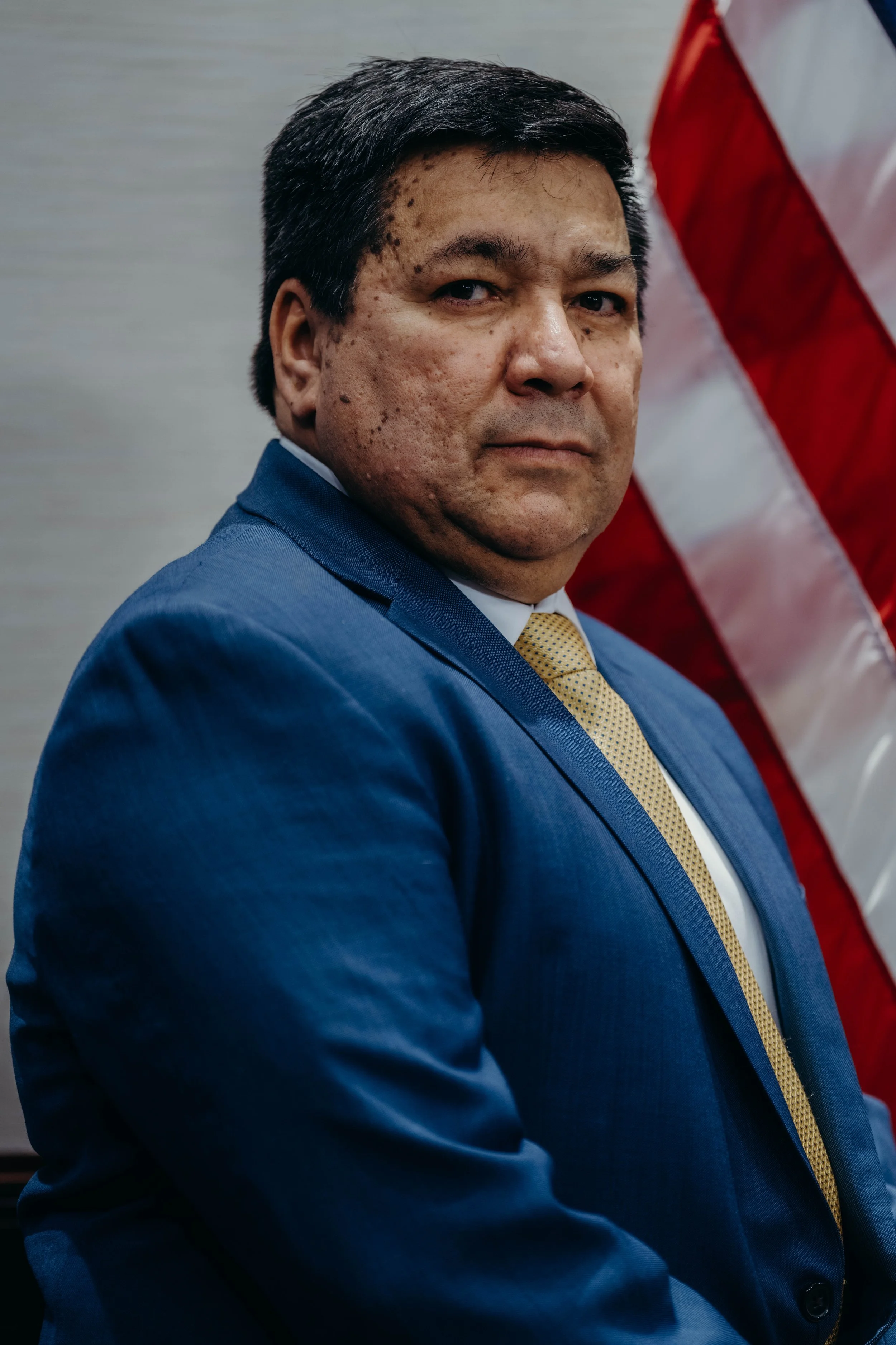 A man in a blue suit with a yellow tie standing in front of an American flag.