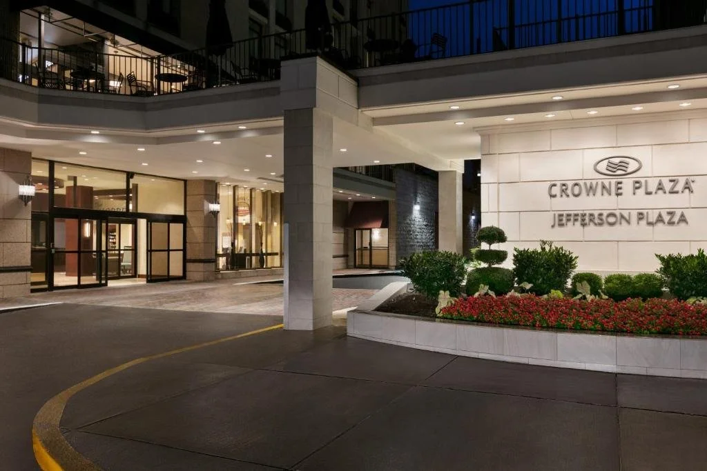 The entrance of Crowne Plaza Jefferson Plaza hotel at night, with a well-lit lobby, glass doors, and landscaped garden with bushes and flowers.